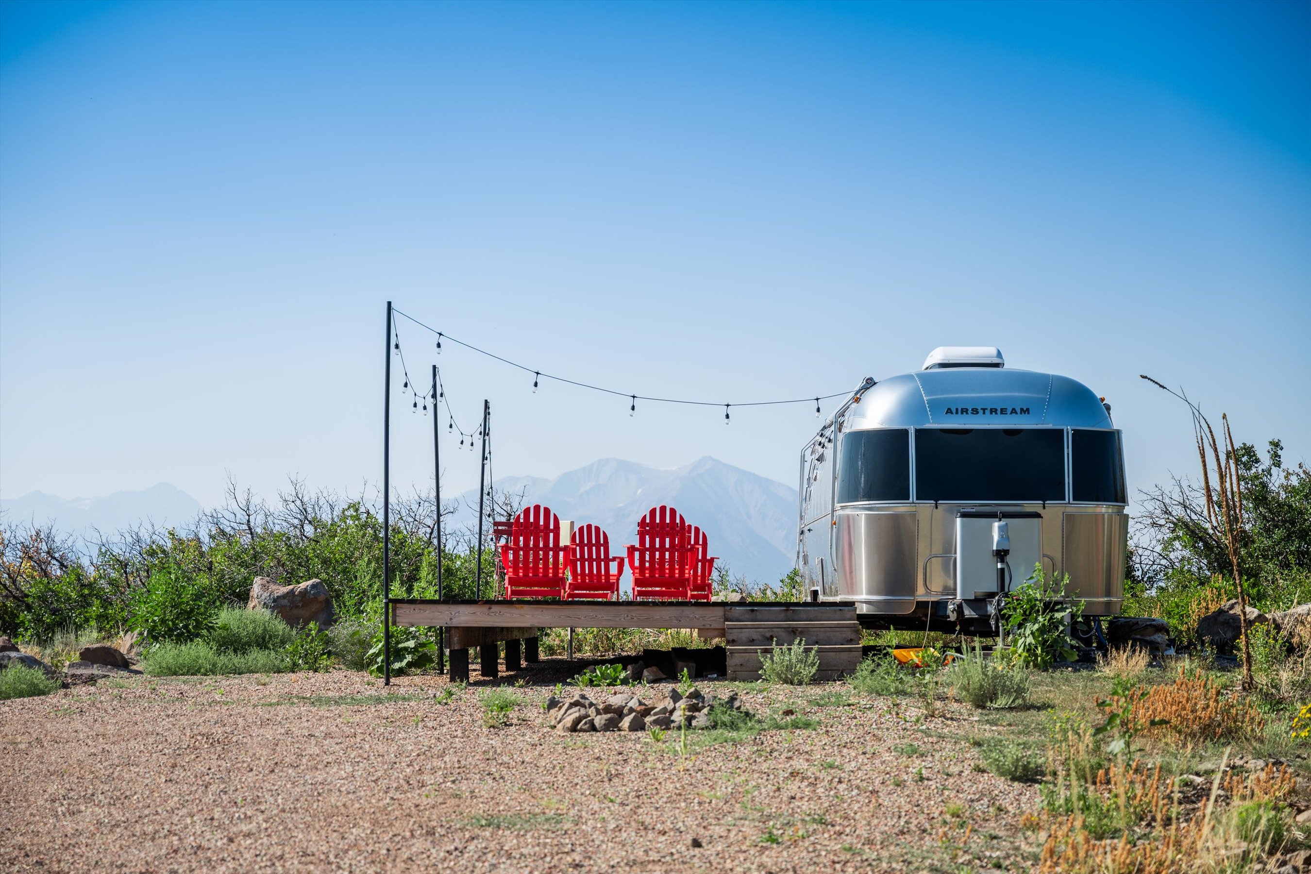 This scenic outdoor space features a wooden deck equipped with three vibrant red Adirondack chairs, perfectly positioned to take in the expansive mountain views. An Airstream trailer is parked adjacent to the deck, which is framed by string lights hanging from tall poles, creating an inviting and rustic glamping atmosphere. The gravel foreground and natural desert landscaping emphasize the serene, remote, and adventurous nature of this unique property.