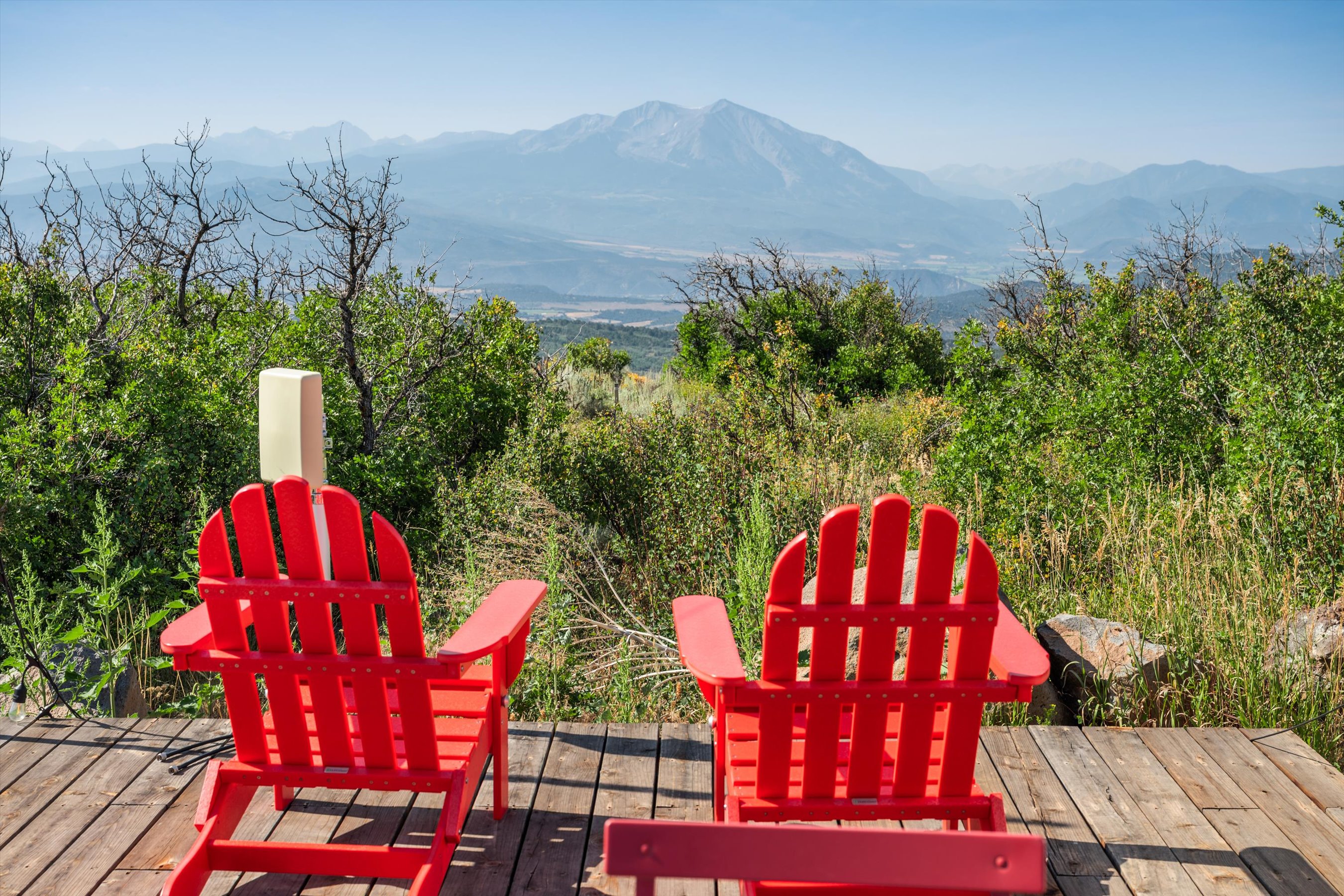 This image captures a serene outdoor deck featuring two vibrant red Adirondack chairs positioned to overlook a breathtaking mountain vista. The wooden deck provides a rustic foundation, while the surrounding lush greenery and expansive valley view create a peaceful, scenic atmosphere perfect for relaxation. The perspective is from behind the chairs, inviting the viewer to imagine themselves sitting and taking in the majestic landscape.