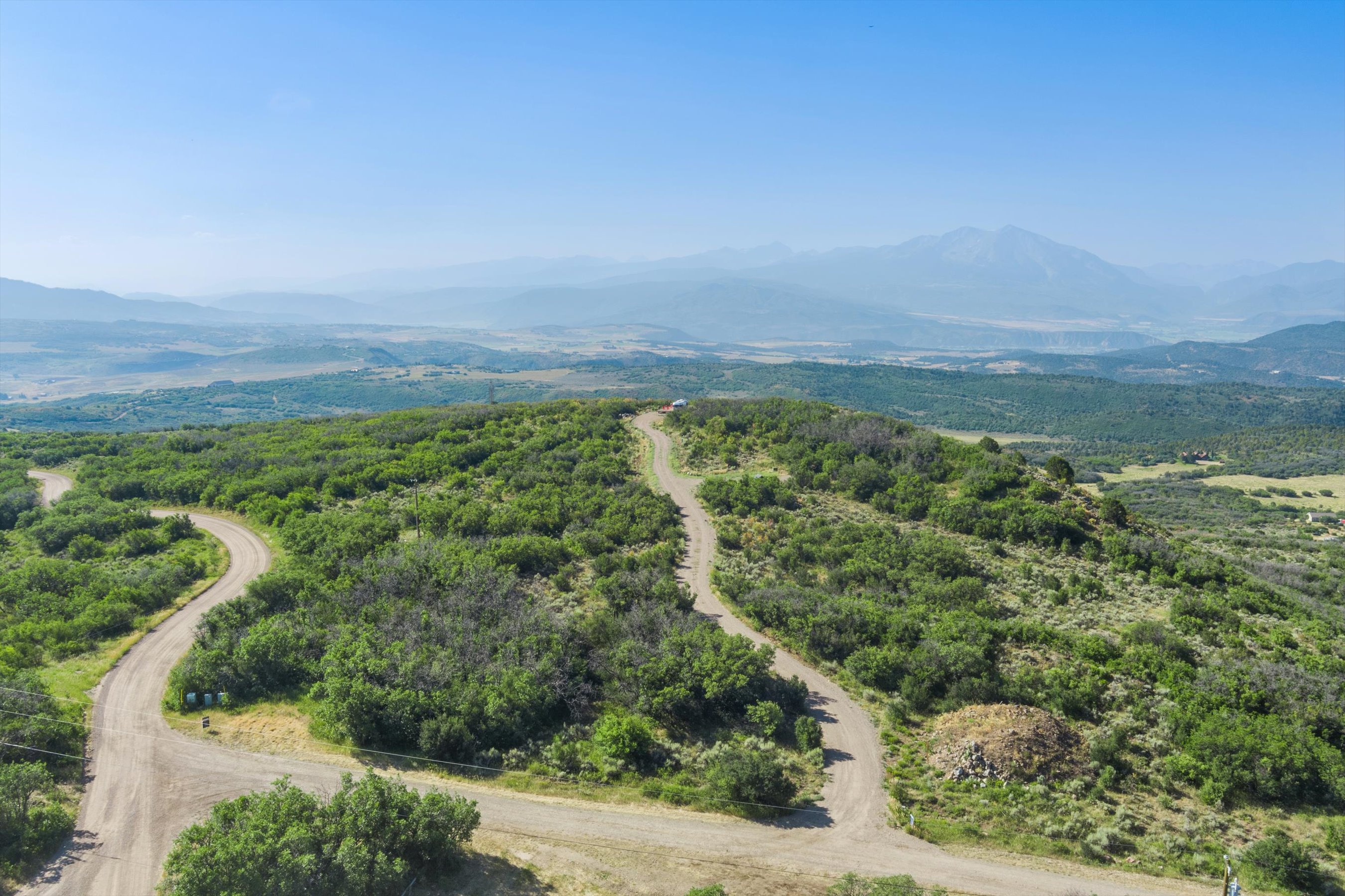 This high-angle aerial view captures a vast, rugged landscape featuring a winding dirt road that cuts through dense, green scrub oak and native vegetation. In the distance, majestic, hazy mountain ranges rise against a clear blue sky, emphasizing the expansive and secluded nature of the property. The perspective provides a comprehensive look at the topography, highlighting the potential for scenic building sites or outdoor recreation.