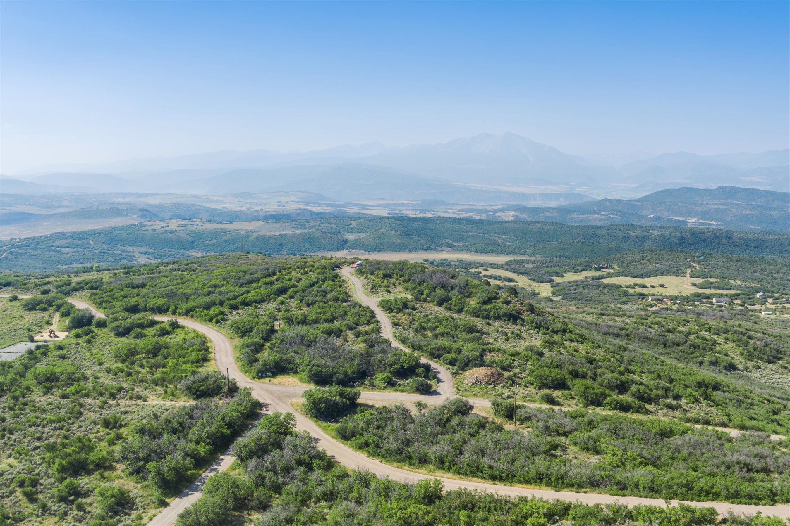 This high-angle aerial view captures a vast, rolling landscape characterized by dense green shrubbery and winding dirt roads that traverse the terrain. In the distance, majestic, hazy mountains rise against a clear blue sky, emphasizing the expansive and secluded nature of the property. The perspective provides a comprehensive look at the topography, suggesting a peaceful, rural setting ideal for those seeking privacy and natural beauty.