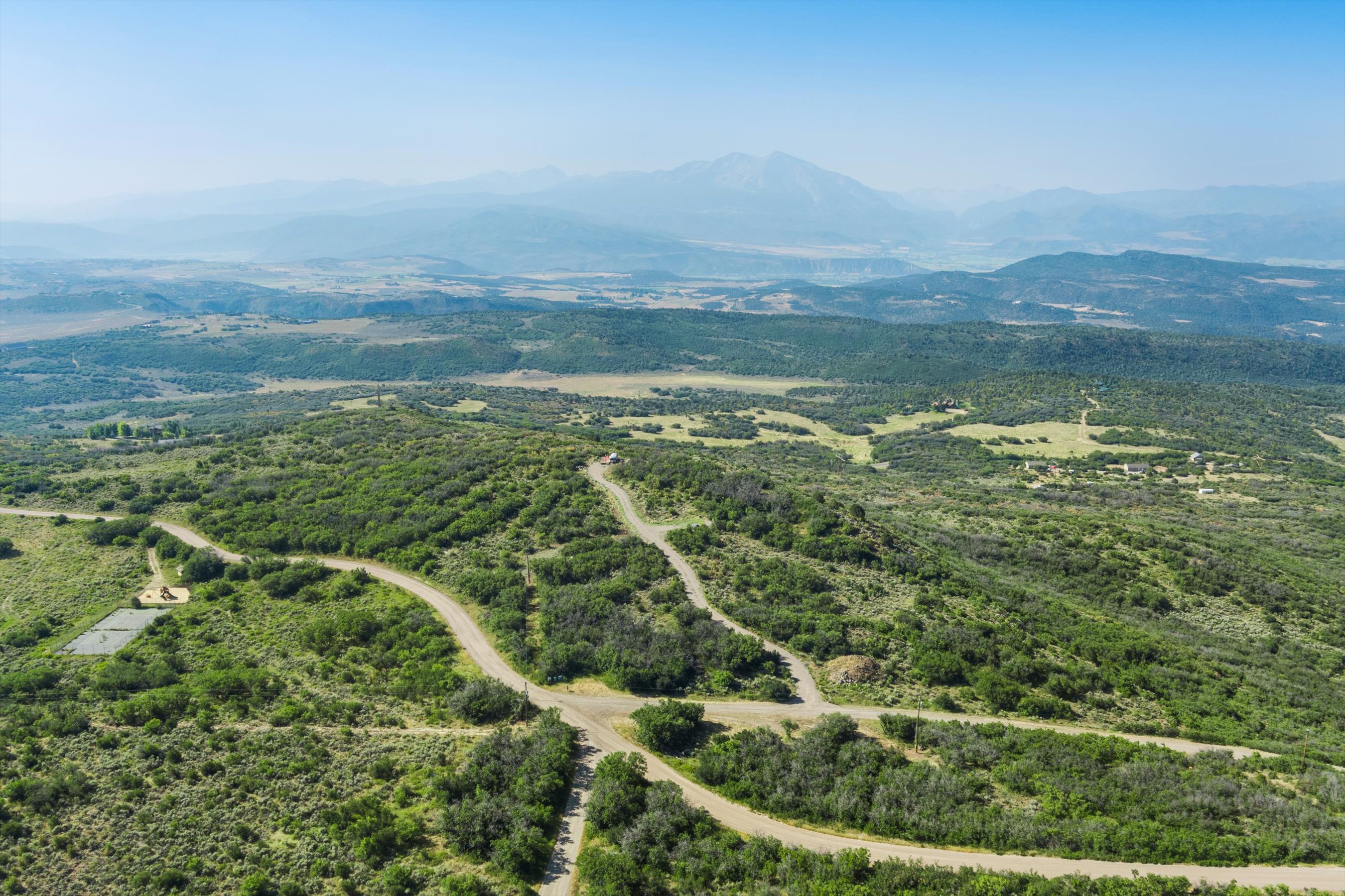 This high-angle aerial view captures a vast, rugged landscape characterized by rolling hills covered in dense green scrub brush and winding dirt roads. In the distance, majestic, hazy mountains rise against a clear sky, emphasizing the expansive and secluded nature of the property. The perspective provides a cinematic sense of scale, highlighting the natural beauty and potential for outdoor exploration in this remote setting.