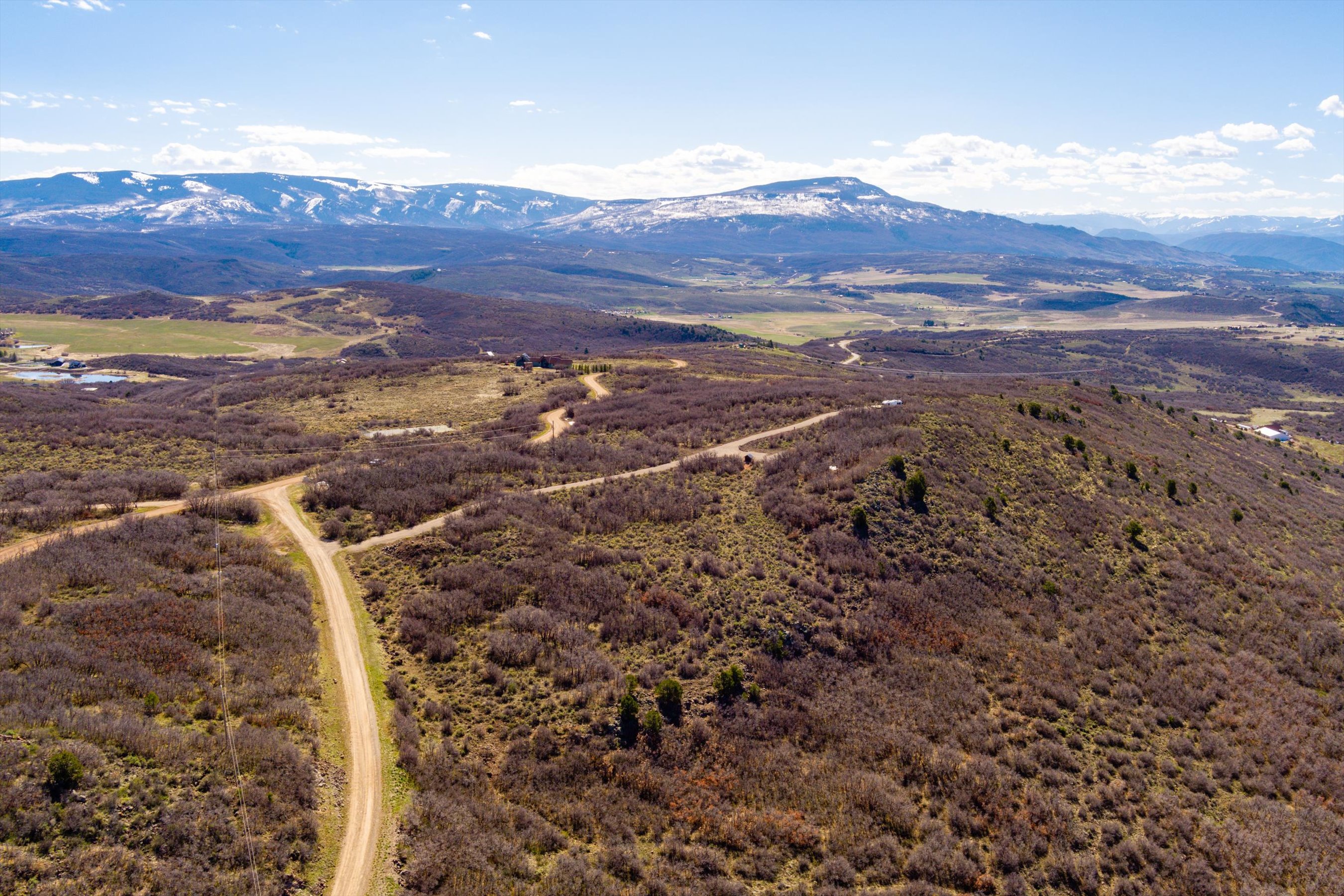 This high-angle aerial shot captures a vast, rugged landscape characterized by rolling hills covered in dense, dormant brush and scrub oak. A winding dirt road cuts through the terrain, leading toward a secluded property nestled on a ridge, with majestic, snow-capped mountains dominating the distant horizon under a clear blue sky. The perspective emphasizes the expansive, private nature of the land and its dramatic topographical features.