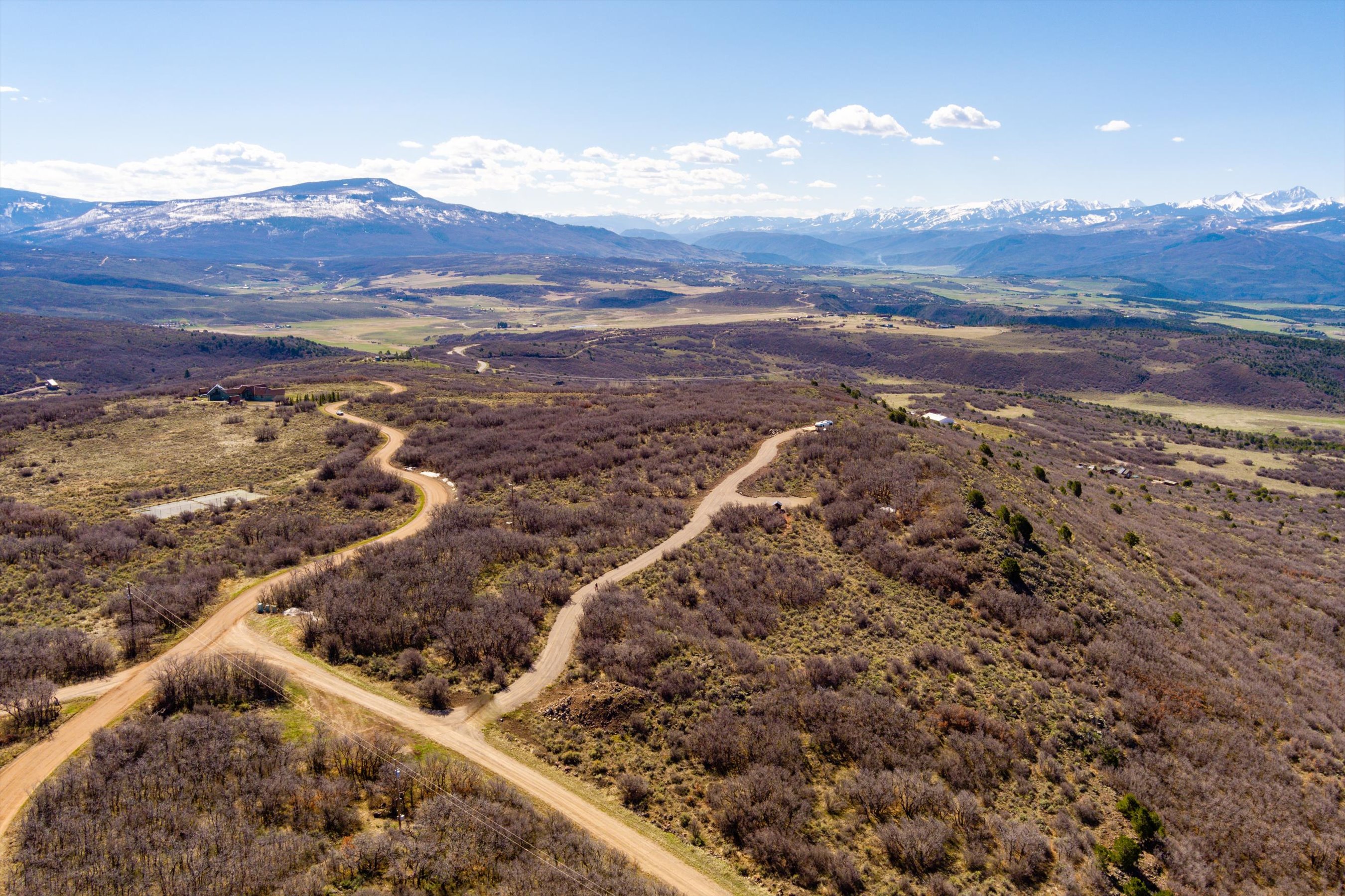 This high-angle aerial shot captures a vast, rugged landscape featuring winding dirt roads that traverse through dense, dormant brush and rolling hills. In the distance, majestic snow-capped mountain ranges rise against a clear blue sky, emphasizing the expansive and secluded nature of the property. The perspective provides a comprehensive overview of the terrain, highlighting the potential for development or private retreat living.