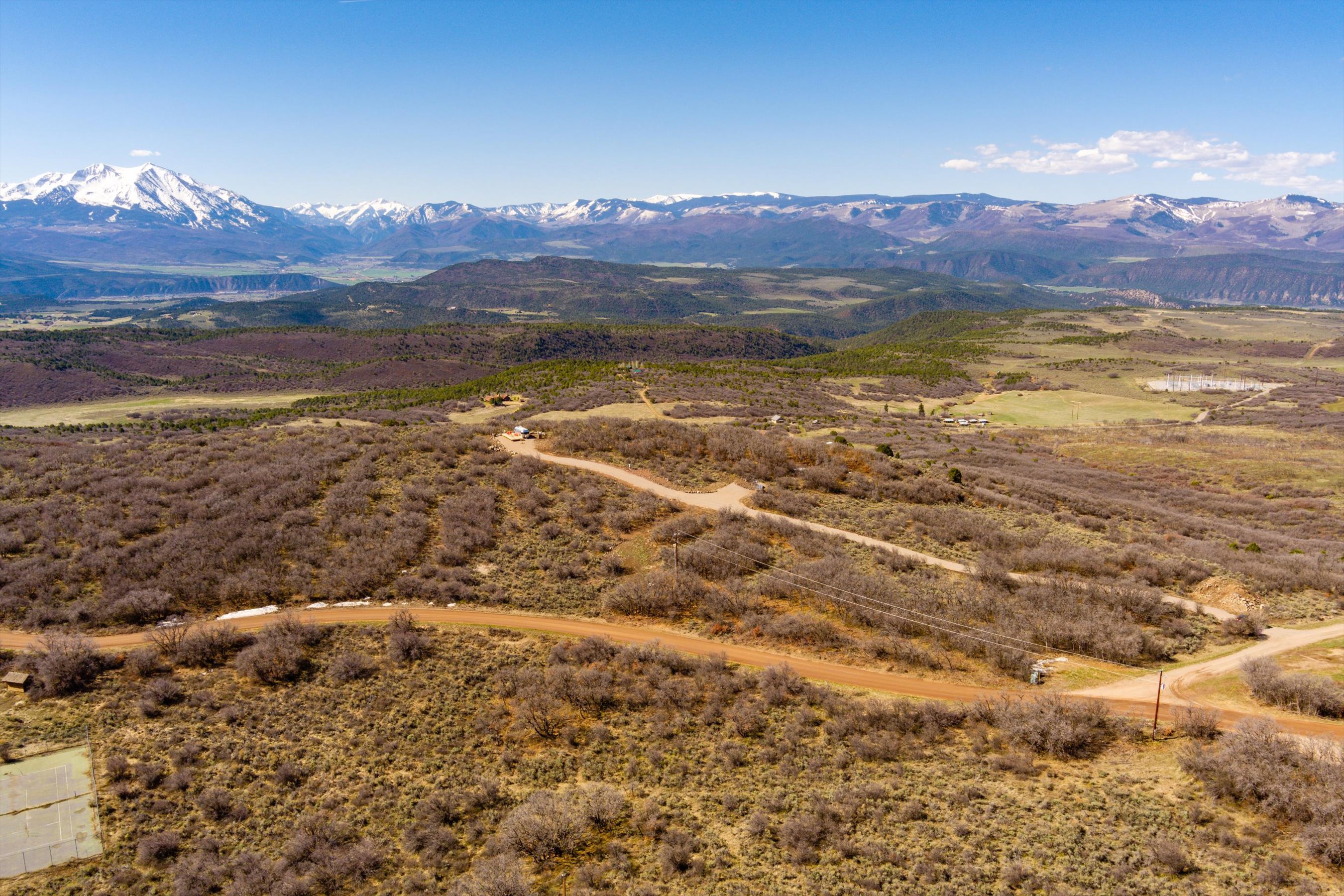 This high-angle aerial shot captures a vast, rugged landscape featuring a winding dirt road that traverses through scrubby, low-lying vegetation. In the distance, a majestic, snow-capped mountain range dominates the horizon under a clear blue sky, emphasizing the property's expansive and scenic rural setting. The perspective highlights the isolation and natural beauty of the terrain, suggesting a peaceful, remote retreat.