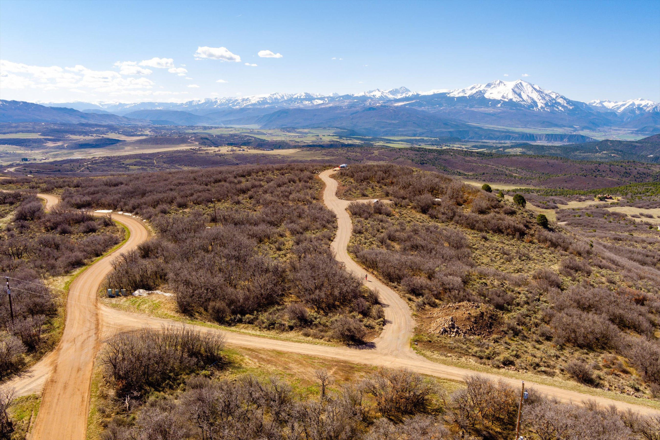 This high-angle aerial view captures a vast, rugged landscape featuring winding dirt roads that traverse through dense, dormant brush and rolling hills. In the distance, a majestic, snow-capped mountain range dominates the horizon under a clear blue sky, emphasizing the property's expansive and secluded natural setting. The perspective highlights the significant acreage and the potential for development or private retreat living in a serene, mountainous environment.