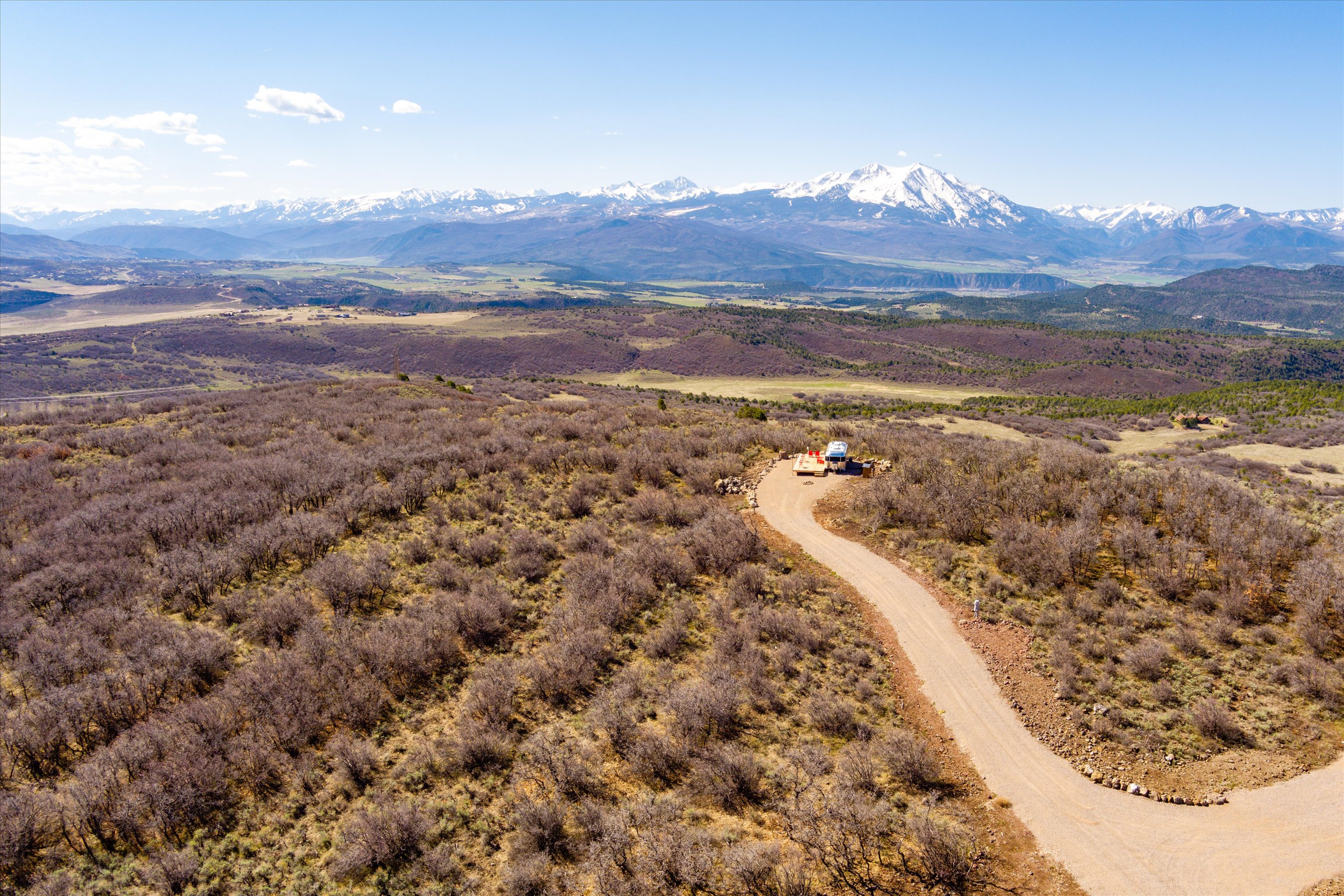 This high-angle aerial shot captures a vast, rugged landscape featuring a winding dirt road leading to a secluded hilltop clearing. A small recreational vehicle is parked on a wooden deck, offering a panoramic view of the expansive valley and the majestic, snow-capped mountain range in the distance. The scene emphasizes the property's remote, serene location and its potential for off-grid living or a private retreat.