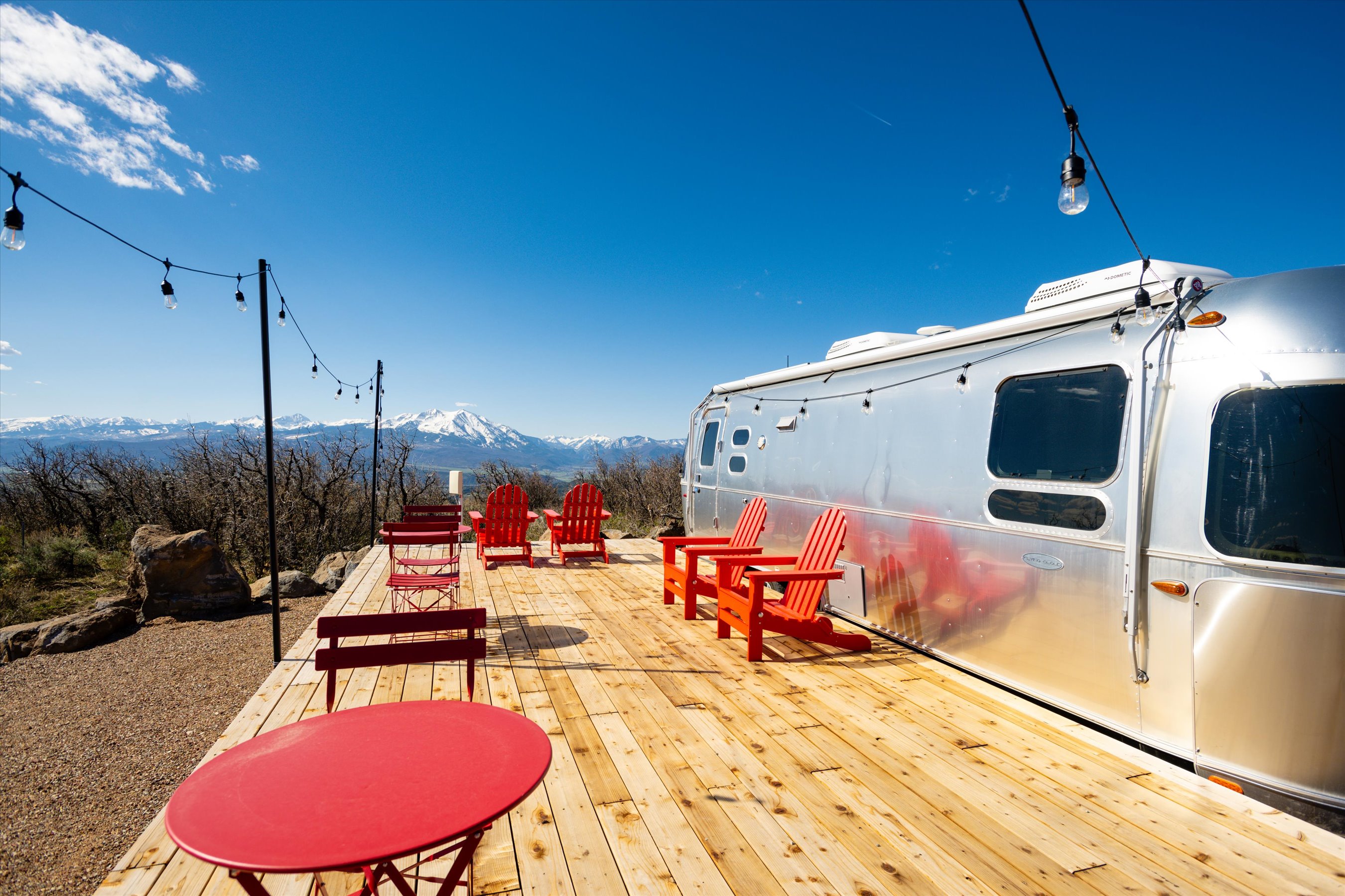 This image showcases a spacious wooden deck featuring a classic Airstream trailer parked alongside several bright red Adirondack chairs and a matching bistro set. The deck offers a breathtaking, unobstructed panoramic view of snow-capped mountains under a clear blue sky, creating an inviting and serene outdoor living space. String lights overhead add a charming, cozy ambiance, perfect for evening relaxation or entertaining in a scenic, high-altitude setting.