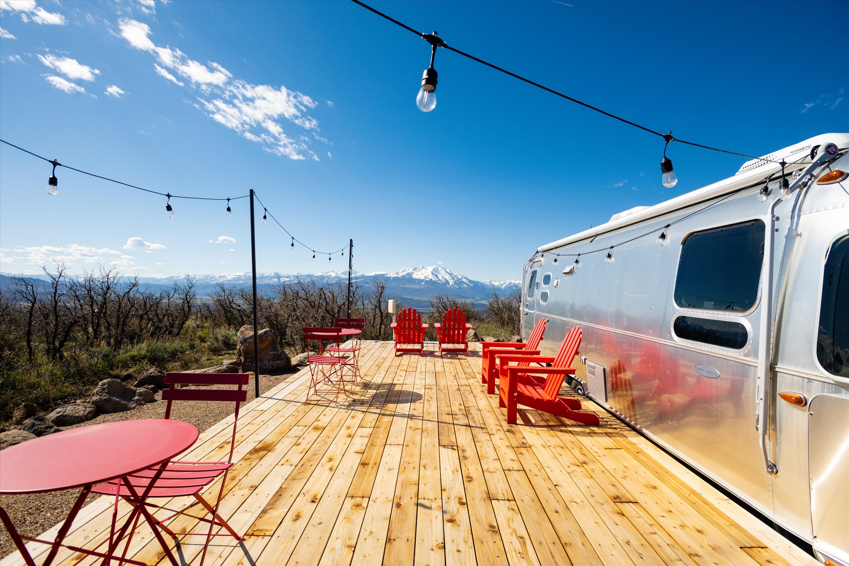 This stunning outdoor deck features a modern Airstream trailer parked alongside vibrant red patio furniture, set against a breathtaking panoramic view of snow-capped mountains. The wooden deck provides a spacious area for relaxation, accented by string lights that create an inviting atmosphere under the clear blue sky. The composition captures a perfect blend of adventurous travel and serene mountain living, ideal for a high-end glamping or vacation rental listing.