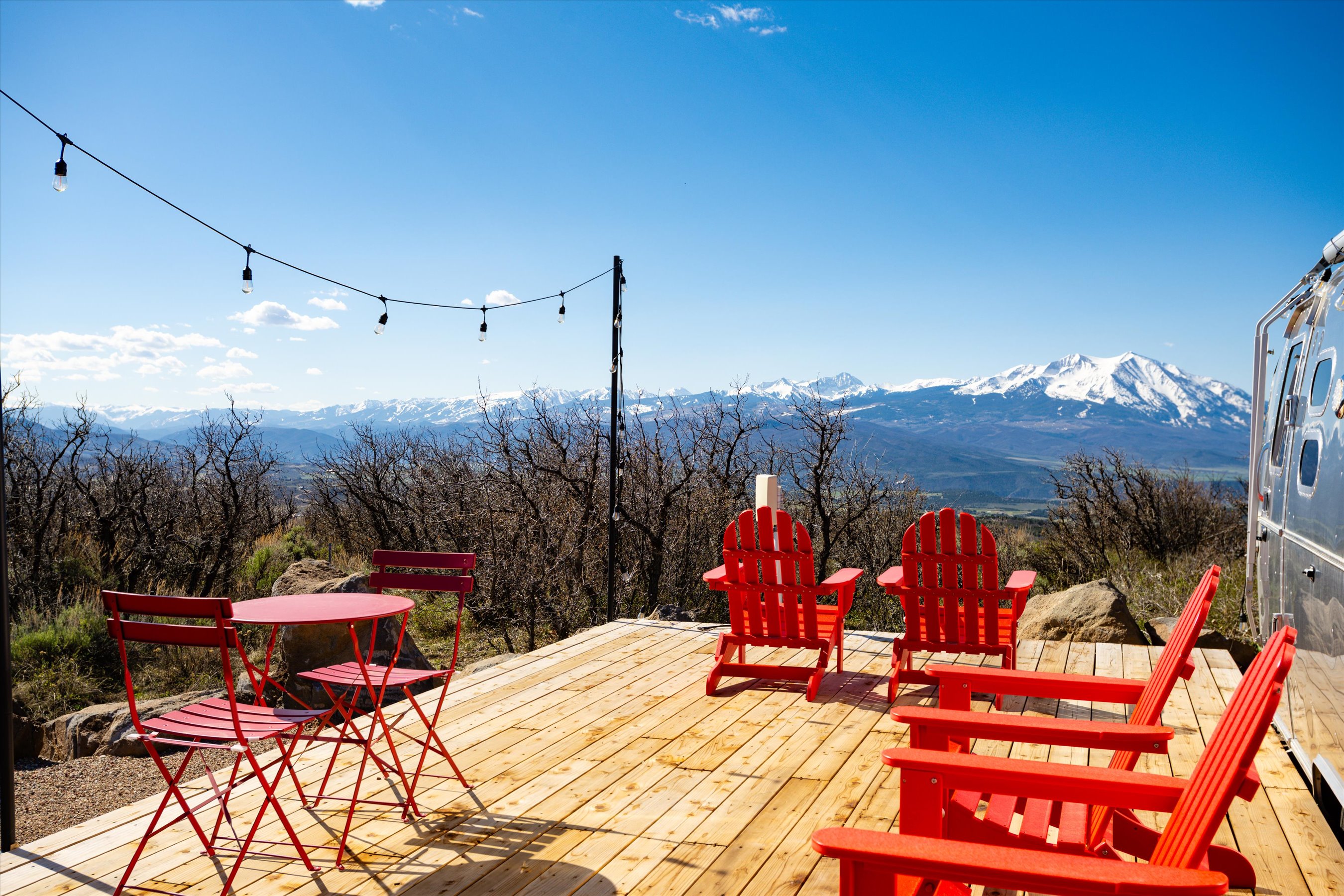 This inviting wooden deck offers a stunning panoramic view of snow-capped mountains, providing an ideal space for outdoor relaxation. The area is furnished with a small bistro set and several vibrant red Adirondack chairs, creating a cozy and picturesque setting. A string of overhead lights adds a charming touch, while the proximity to a sleek Airstream trailer enhances the adventurous, glamping-inspired aesthetic.