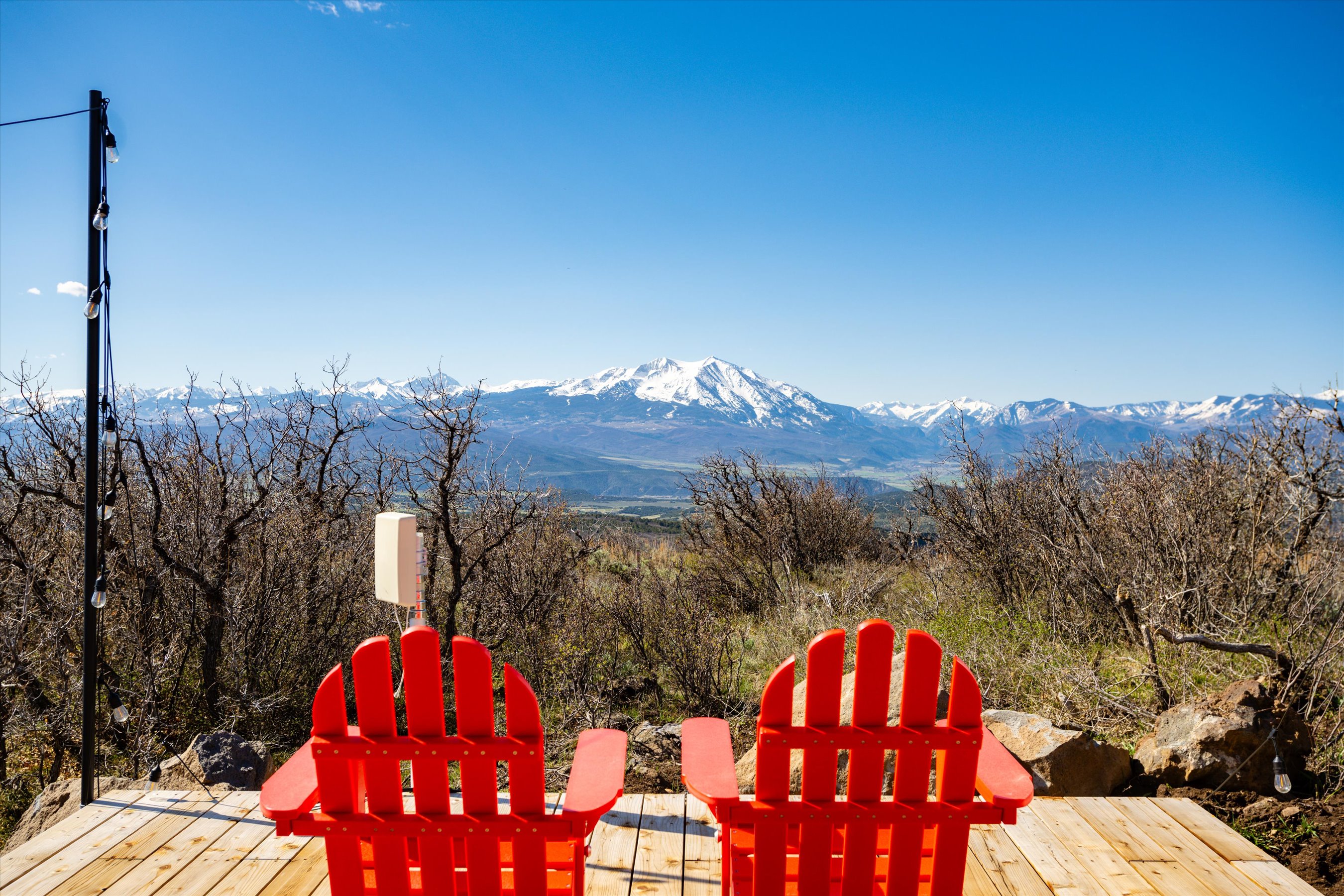 This image captures a serene outdoor deck featuring two vibrant red Adirondack chairs positioned to overlook a breathtaking mountain range. The wooden deck provides a rustic platform, while a string of lights on a pole adds a touch of charm to the expansive, clear-sky landscape. The perspective is from behind the chairs, inviting the viewer to imagine sitting down and taking in the panoramic, scenic vista.