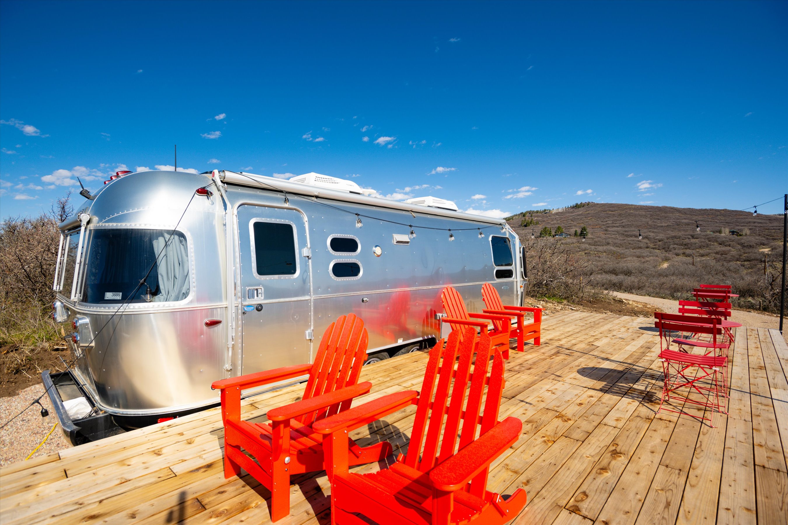 This image showcases a modern, silver Airstream trailer parked on a spacious wooden deck, creating a stylish glamping retreat. The deck is furnished with several vibrant red Adirondack chairs and a small bistro set, perfect for outdoor relaxation while enjoying the expansive, rugged landscape in the background. The bright, clear blue sky and natural surroundings emphasize a serene and adventurous outdoor living experience.