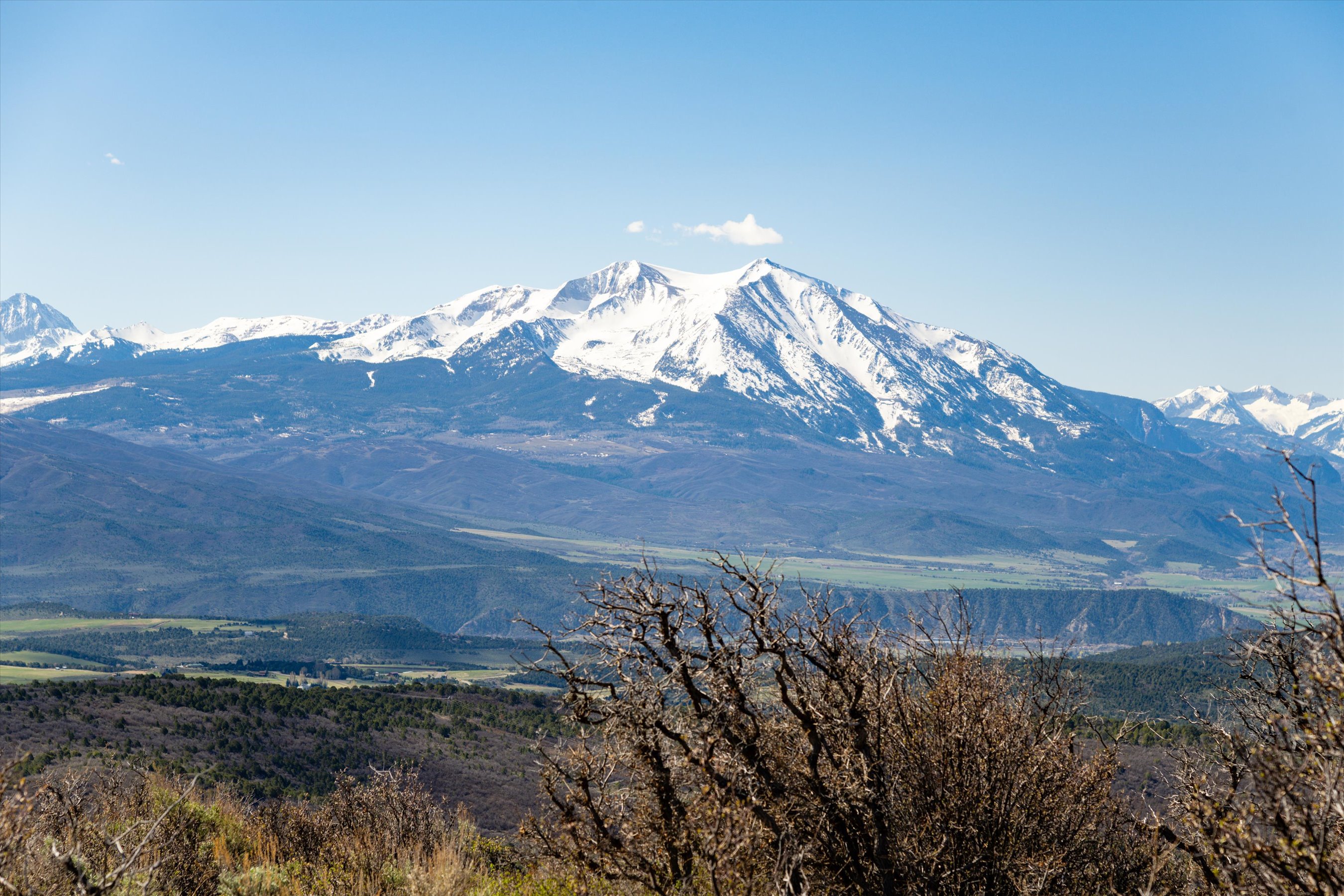 This wide-angle landscape shot captures a majestic, snow-capped mountain range rising above a sprawling valley. In the foreground, dry, leafless shrubs and brush frame the view, emphasizing the vast scale and natural beauty of the surrounding terrain. The clear blue sky and expansive vistas suggest a serene, high-altitude location ideal for those seeking privacy and connection with nature.