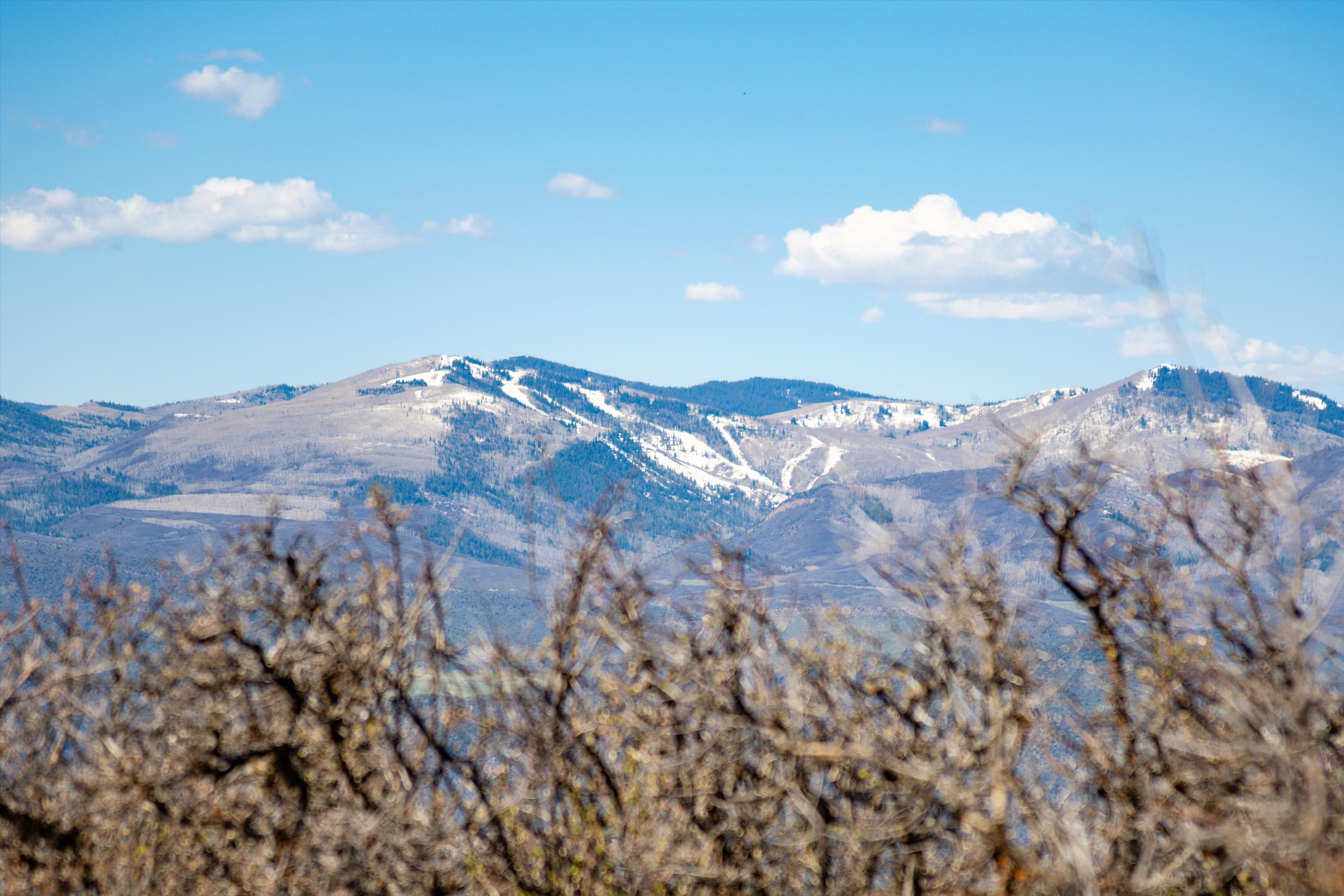 This image captures a scenic, long-range view of a mountain range with patches of snow still visible on the slopes, suggesting a transition between seasons. In the foreground, out-of-focus, leafless branches frame the landscape, adding depth and a natural, rustic texture to the composition. The clear blue sky and bright lighting emphasize the expansive, tranquil nature of the mountain vista, making it an appealing feature for a property with a view.