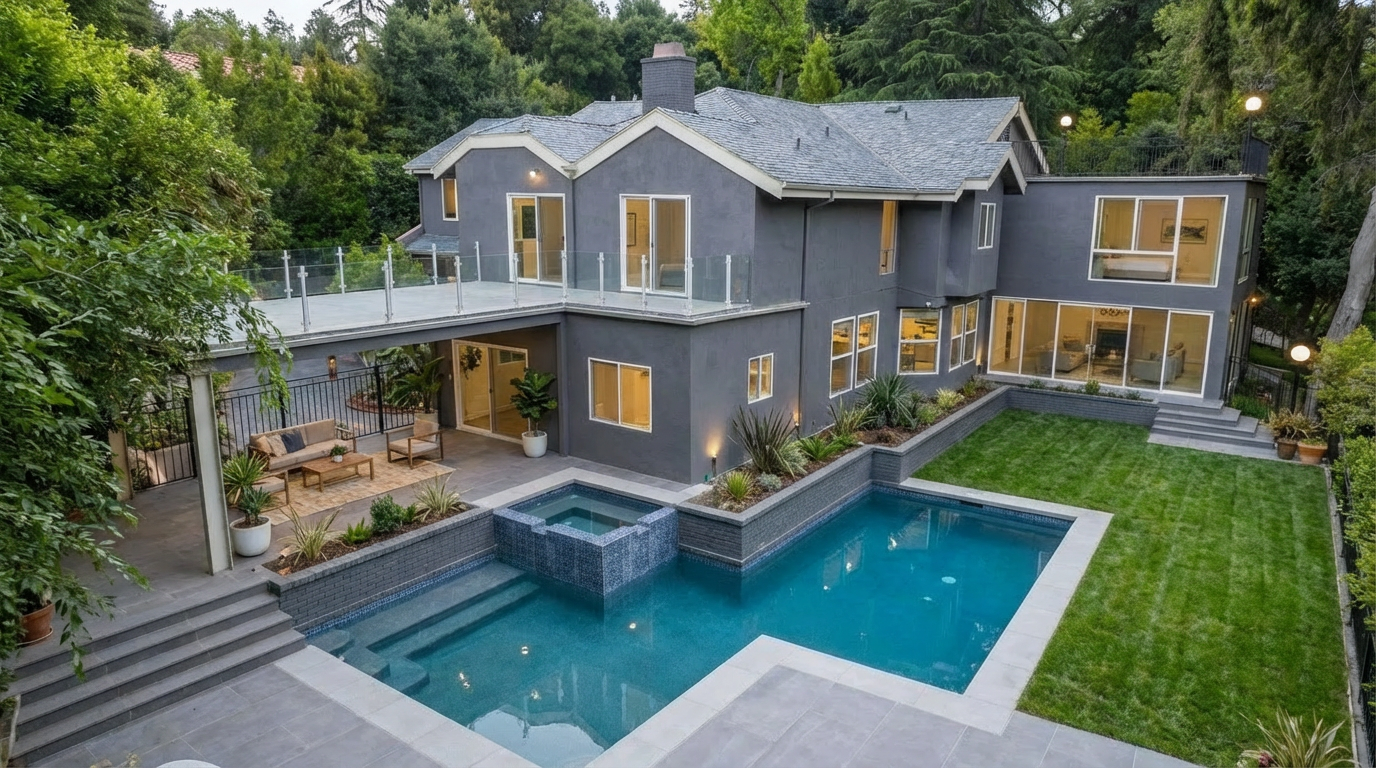 This image showcases the rear exterior of a modern home, emphasizing the luxurious pool and spa area. The pool features a unique geometric design with integrated spa, surrounded by a well-manicured lawn and patio space. A covered outdoor lounge area with stylish furniture is accessible from an upper-level deck, creating an inviting space for relaxation and entertainment.