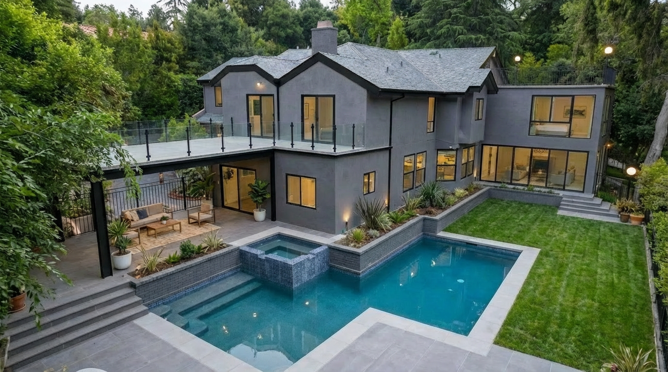 This image showcases the rear exterior of a modern home, emphasizing the luxurious pool and spa area. The pool features a unique design with integrated spa and steps, surrounded by well-manicured lawn and landscaping. A covered patio with outdoor seating extends from the house, offering a relaxing space overlooking the pool area.
