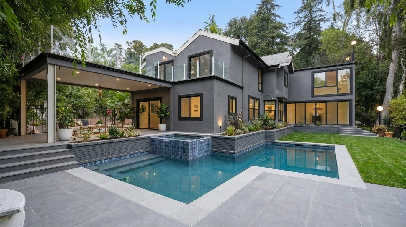 This image showcases the rear exterior of a modern home, highlighting a luxurious pool and spa area. The pool features a clean, geometric design with light-colored coping, while the adjacent spa is tiled with blue mosaics. The home's architecture includes a covered patio area and multiple windows, creating a seamless indoor-outdoor living experience.