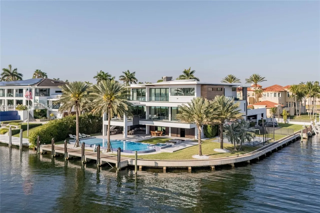 This aerial view showcases a luxurious waterfront property featuring a modern, multi-story home with expansive windows and balconies. A large swimming pool and well-manicured lawn extend to the water's edge, complemented by a private dock. Palm trees dot the landscape, enhancing the tropical ambiance and highlighting the property's prime location.
