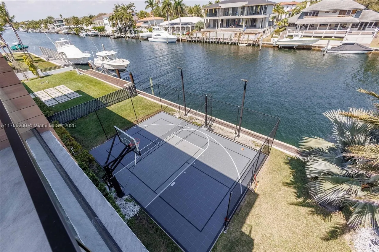 This aerial view showcases a waterfront property featuring a modern basketball court enclosed by fencing, situated adjacent to a canal with boats docked nearby. The property includes well-maintained landscaping and contemporary architectural elements, creating an appealing outdoor recreational space. The perspective is from above, providing a comprehensive view of the property's layout and its integration with the waterfront environment.