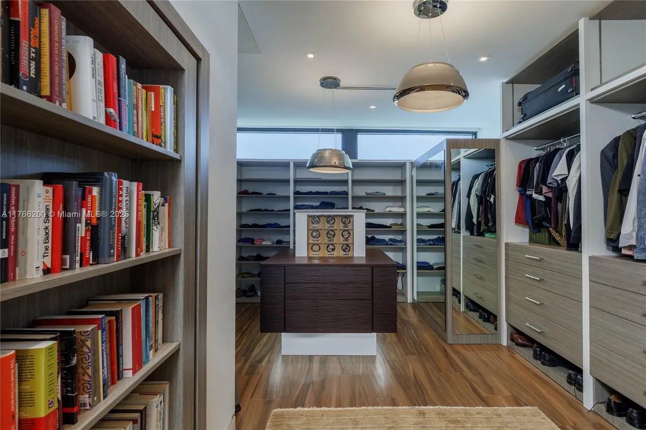 This is a well-organized walk-in closet featuring custom shelving, drawers, and hanging space. The closet includes a central island with drawers and a display case, hardwood flooring, and modern pendant lighting. A bookshelf is visible on the left, adding a touch of sophistication to the space.