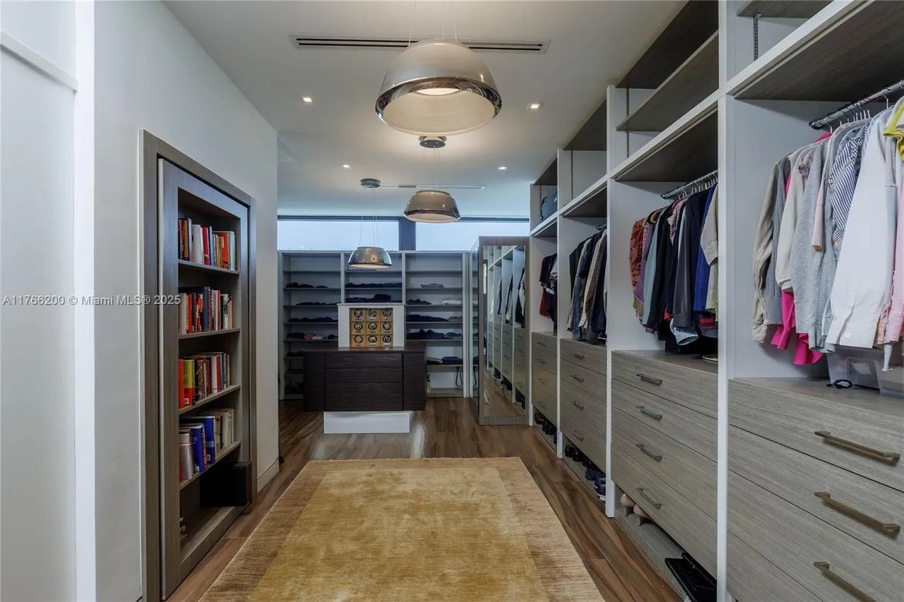 This is a well-organized walk-in closet featuring custom shelving and drawers. The closet is illuminated by modern pendant lights and recessed lighting. A built-in bookcase adds a unique touch, and a large rug anchors the space, creating a luxurious and functional dressing area.