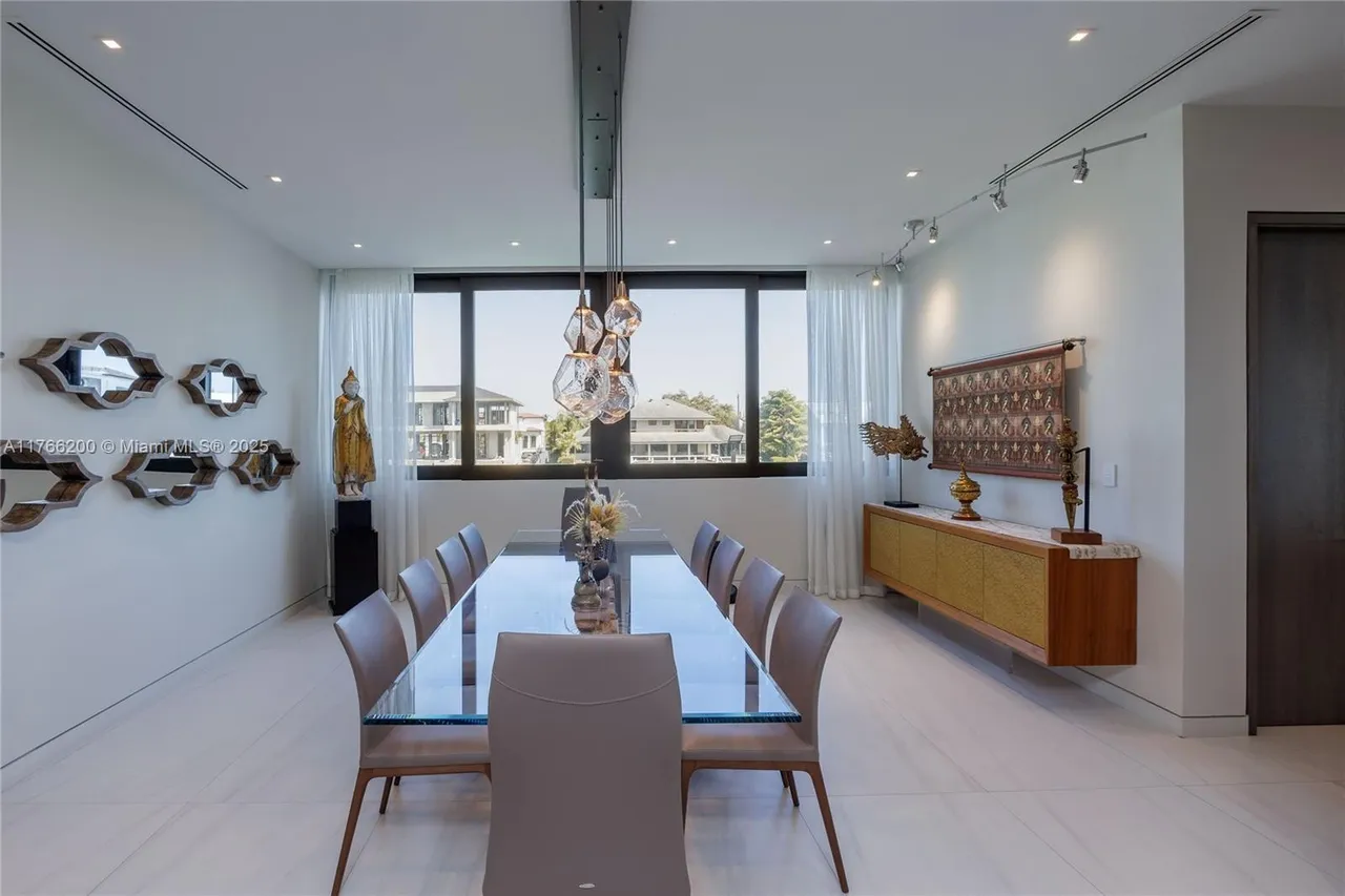 This is a bright and modern dining room featuring a long glass-topped table with seating for ten. The room is decorated with contemporary art and sculptures, and the large windows offer a view of the outdoors. The overall impression is one of elegance and sophistication.