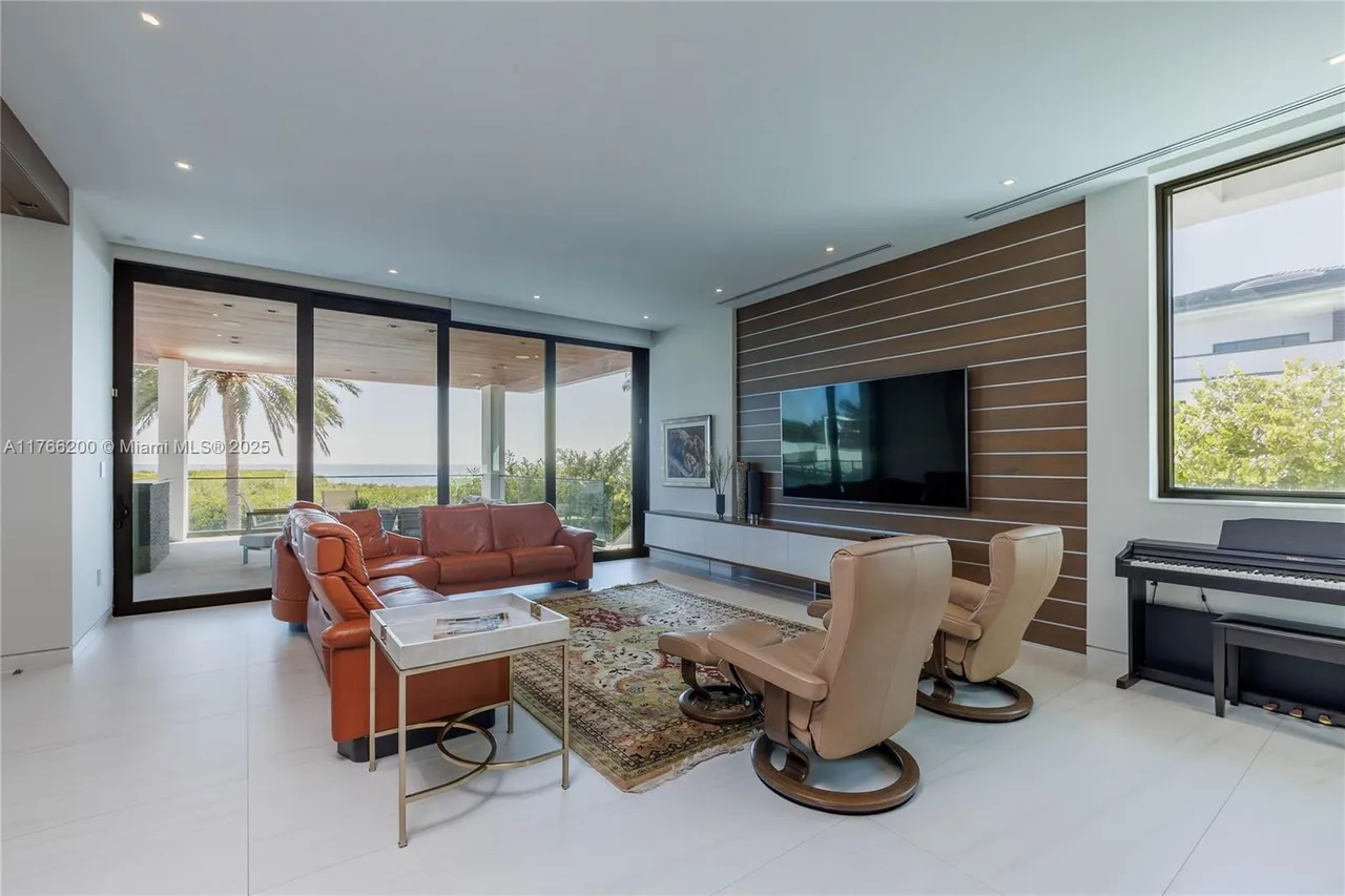 This is a spacious living room featuring a large leather sectional sofa, two swivel chairs, and a modern entertainment center with a flat-screen TV. The room has large windows offering a view of the ocean, and the flooring is light-colored tile. The overall style is contemporary and luxurious, creating a comfortable and inviting space.