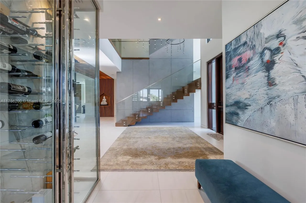 This interior shot showcases a modern hallway with a striking staircase featuring wooden steps and a glass railing. A large abstract painting adorns the wall, complemented by a blue bench. A glass-enclosed wine cellar is visible on the left, adding a touch of luxury to the space.