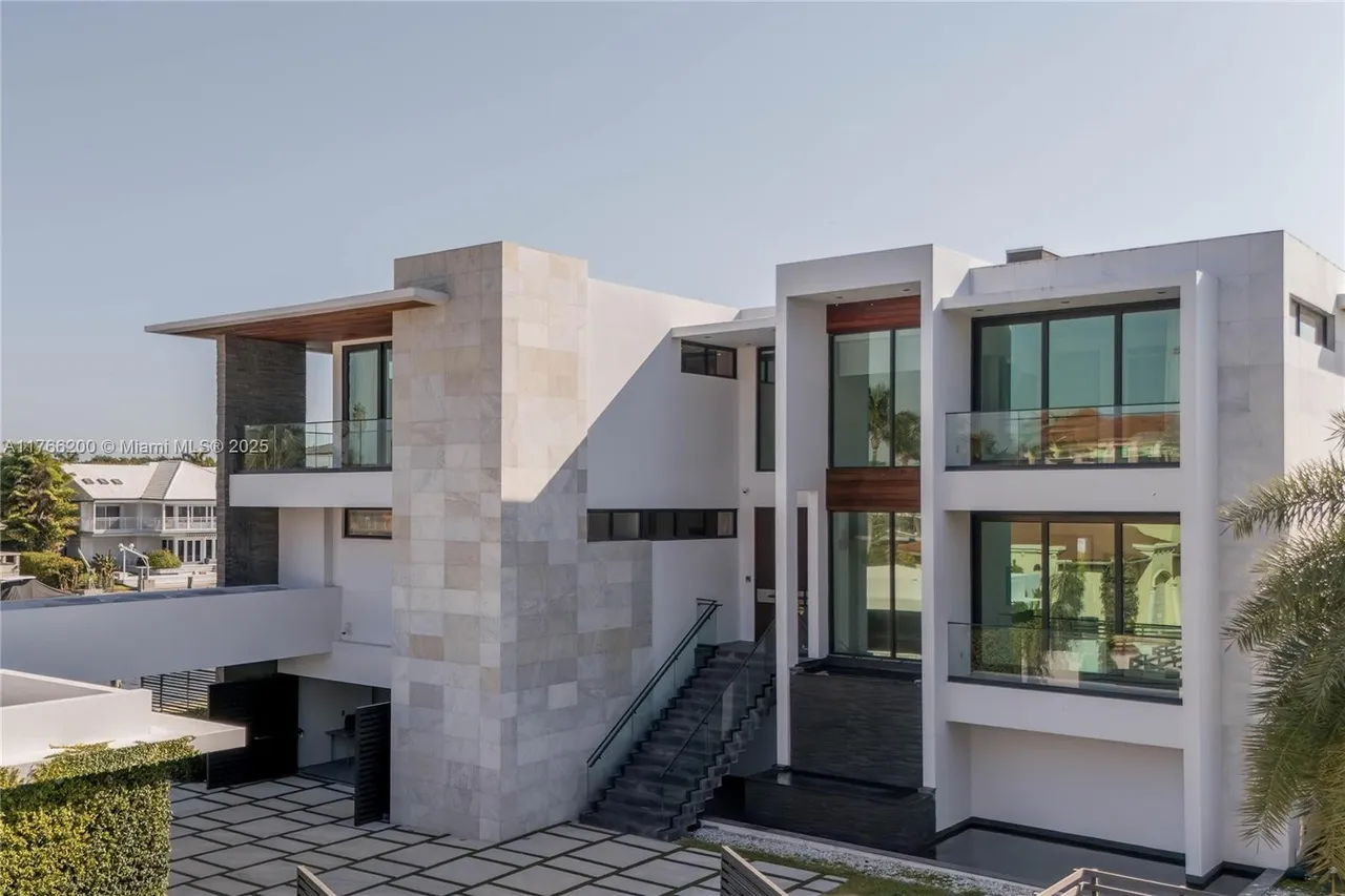 This is a front exterior view of a modern, multi-story home. The architectural style features a combination of white walls, stone accents, and large windows with glass railings. The property includes a paved driveway and landscaping, creating a luxurious and contemporary impression.