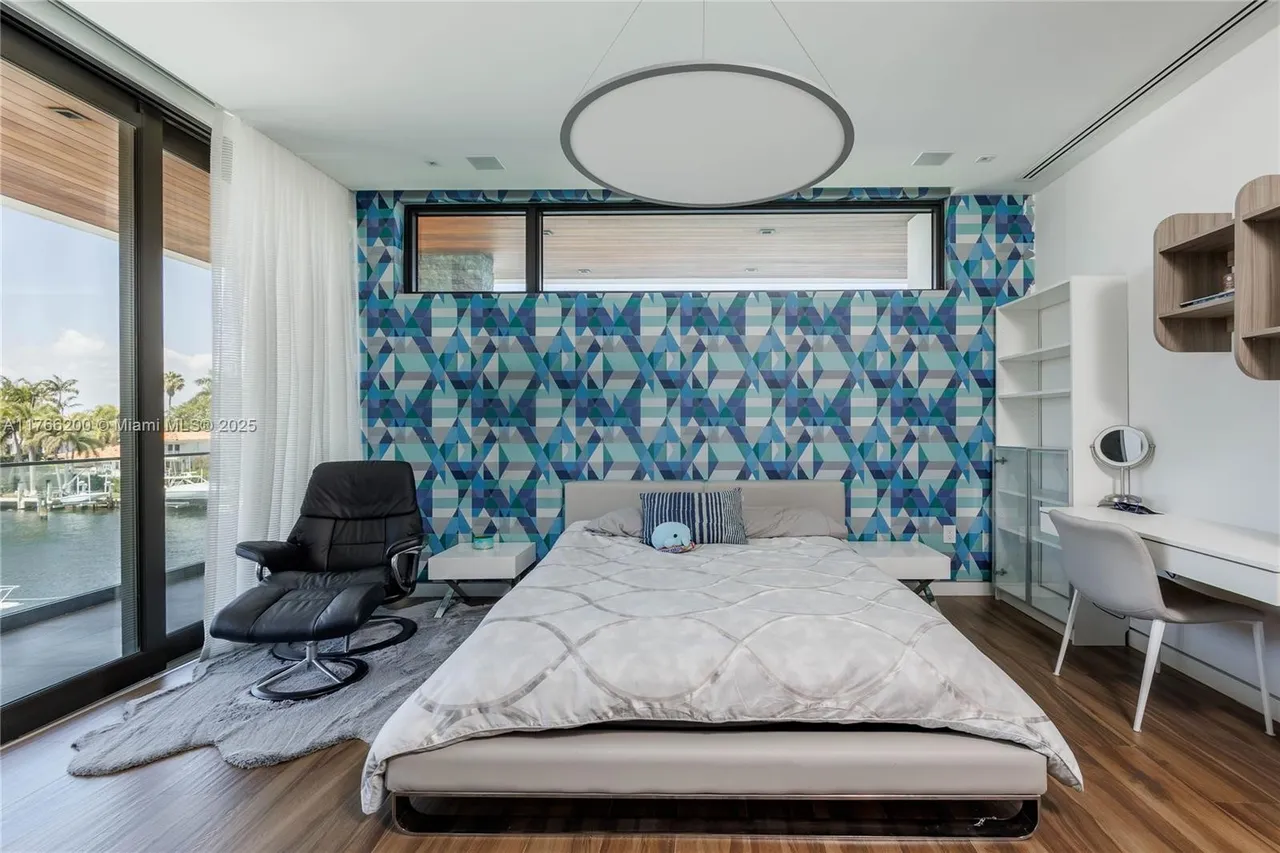 This is a modern bedroom featuring a geometric patterned accent wall in shades of blue and gray. The room includes a platform bed with a light gray comforter, a black leather lounge chair, and a white desk area with shelving. A large window provides natural light and a view of the waterfront, enhancing the room's contemporary and serene atmosphere.