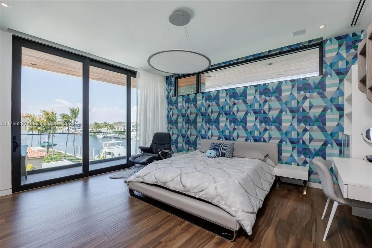 This primary bedroom features a modern design with a geometric patterned accent wall and a large sliding glass door leading to a balcony with water views. The room is furnished with a platform bed, a black leather lounge chair, and a white desk area, complemented by hardwood flooring and a unique circular pendant light. The overall impression is luxurious and contemporary.