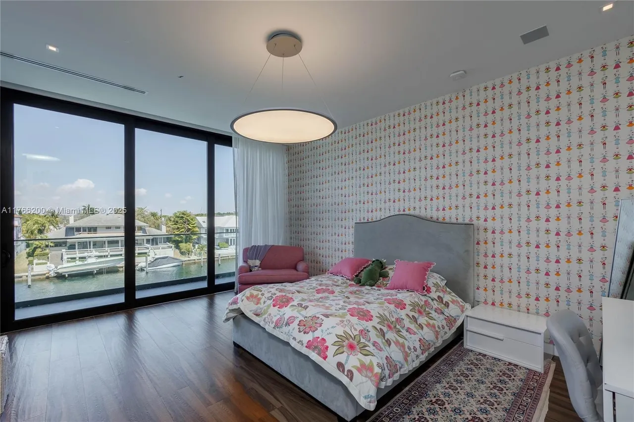 This is a charming bedroom featuring a bed with floral bedding, pink pillows, and a stuffed dinosaur. The room has a unique wallpaper with a doll pattern and a large window offering a waterfront view. The dark wood flooring and modern lighting fixture add a touch of elegance to the space.