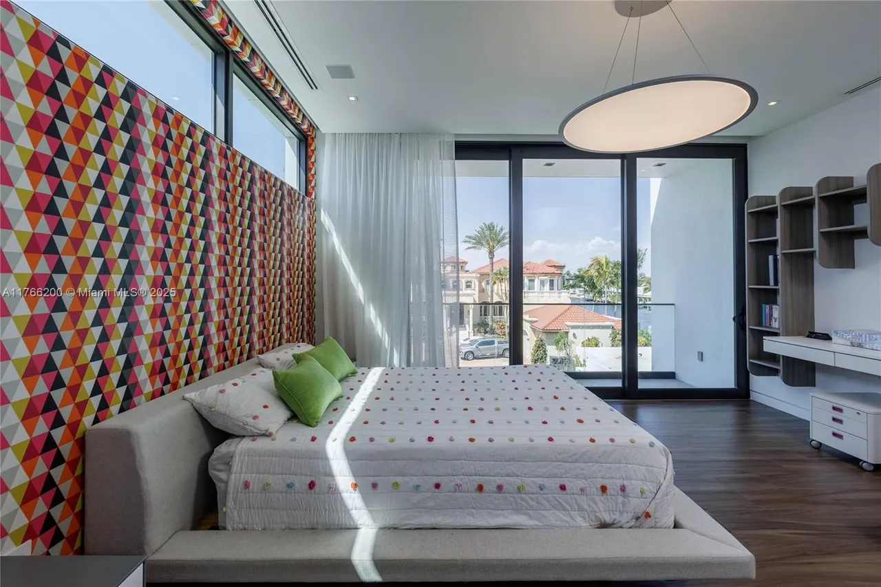 This is a primary bedroom featuring a large bed with a patterned headboard and a white comforter with colorful accents. The room has large windows offering a view of the outdoors, and a modern light fixture hangs from the ceiling. A built-in shelving unit and desk area are visible on the right side of the room, adding functionality to the space.