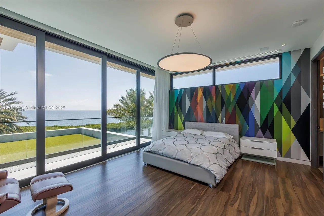 This is a primary bedroom featuring floor-to-ceiling windows with ocean views, a modern bed with a patterned duvet, and contemporary nightstands. The room is decorated with a bold, geometric accent wall and a unique circular pendant light. The hardwood floors add warmth to the space, creating a luxurious and inviting atmosphere.