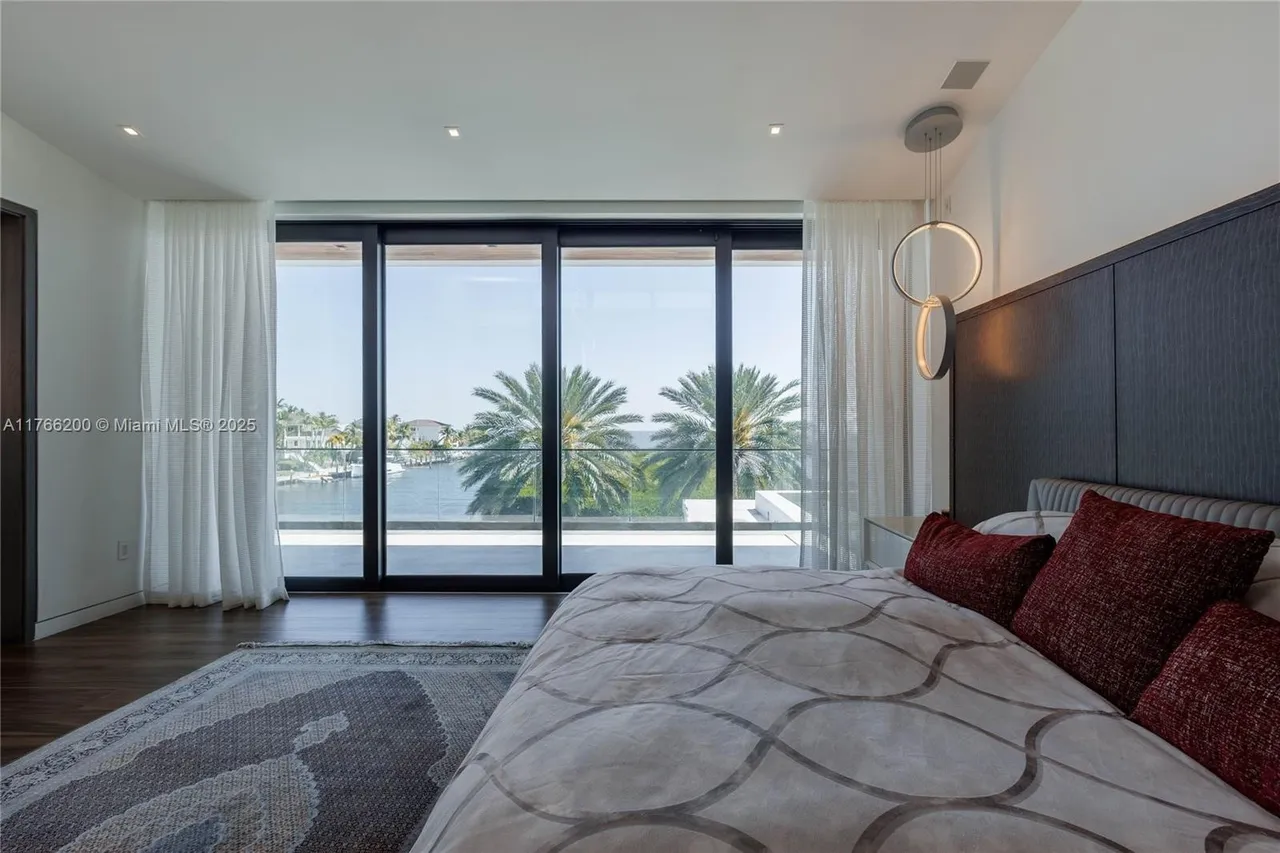 This is a primary bedroom featuring a large bed with patterned bedding and red pillows, a dark headboard, and a modern circular pendant light. The room has floor-to-ceiling windows offering a view of palm trees and the waterfront, with sheer curtains adding a touch of elegance. A gray rug is partially visible in the foreground, adding texture to the dark wood flooring.
