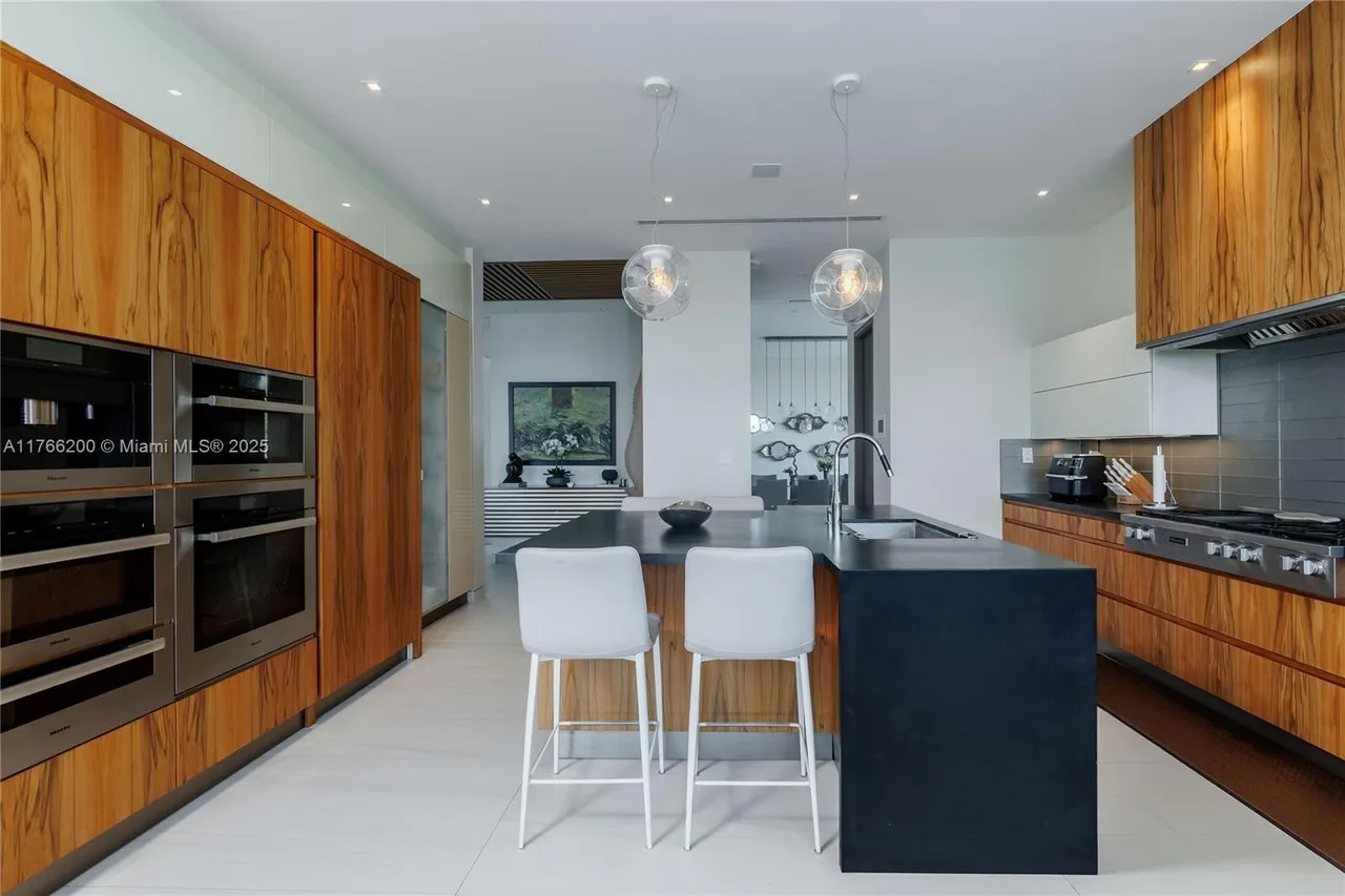 This is a modern kitchen featuring wood cabinetry and a dark countertop island with seating. Stainless steel appliances are built into the cabinetry, and pendant lights hang above the island. The kitchen has a clean and contemporary style with an open layout.