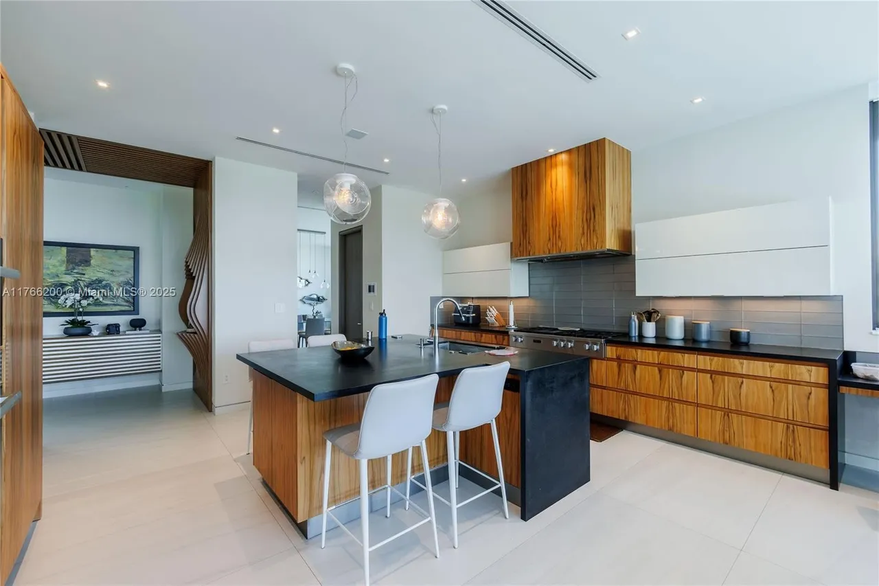 This is a modern kitchen featuring wood cabinetry, a dark countertop island with bar seating, and pendant lighting. The kitchen has a sleek design with a combination of wood and white cabinets, stainless steel appliances, and a gray backsplash. The space is open and well-lit, creating a contemporary and inviting atmosphere.