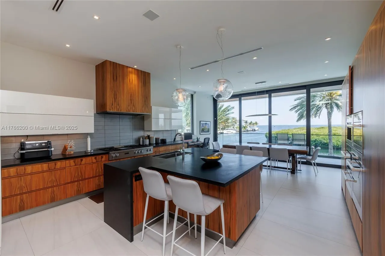 This is a modern kitchen with wood and white cabinetry, a black countertop island with seating, and stainless steel appliances. Large windows offer a view of the water and palm trees, bringing natural light into the space. The kitchen seamlessly transitions into a dining area, creating an open and airy atmosphere.