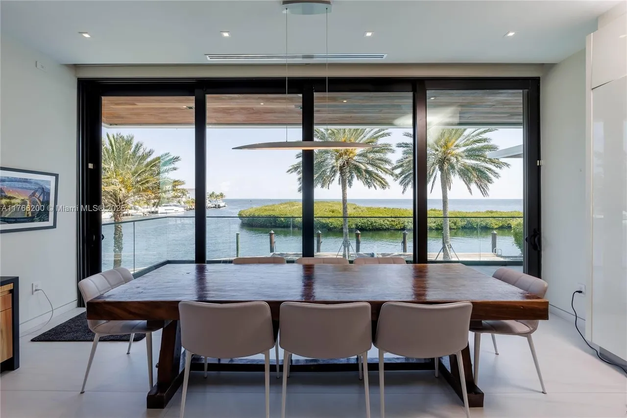 This is an interior shot of a dining room featuring a large, dark wood dining table surrounded by modern chairs with light upholstery. The room has floor-to-ceiling windows offering a stunning view of the water, palm trees, and a small island. A modern, linear pendant light hangs above the table, adding to the contemporary aesthetic of the space.