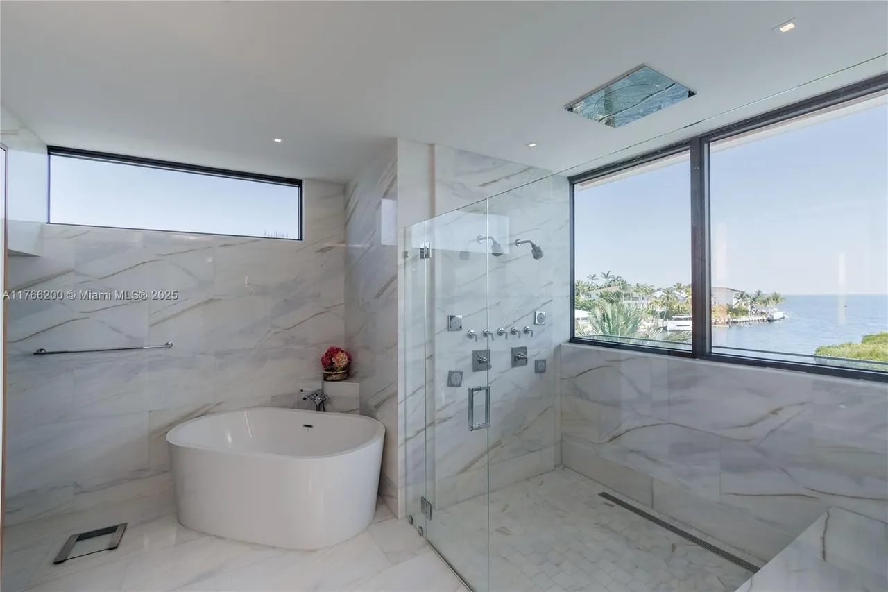 This is a luxurious primary bathroom featuring marble walls and flooring, a freestanding soaking tub, and a glass-enclosed shower with multiple showerheads. A large window provides natural light and a view of the water. The bathroom exudes a spa-like atmosphere with its clean lines and high-end finishes.