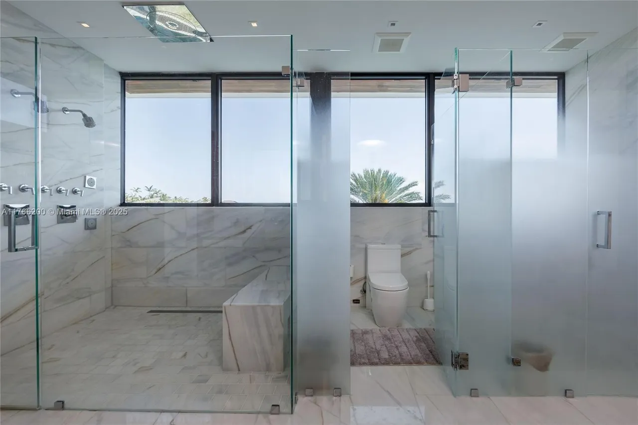 This is a luxurious primary bathroom featuring a glass-enclosed shower with multiple showerheads and a built-in bench. The walls and floors are covered in marble, creating a clean and elegant aesthetic. A separate toilet area is enclosed with frosted glass, providing privacy.