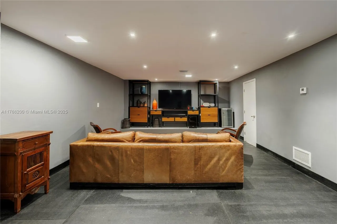 This is an interior shot of a living room featuring a large, tan leather sofa as the focal point. Behind the sofa, there's a media center with a flat-screen TV flanked by shelving units. The room has a modern, minimalist aesthetic with gray walls and dark flooring, creating a cozy yet contemporary atmosphere.