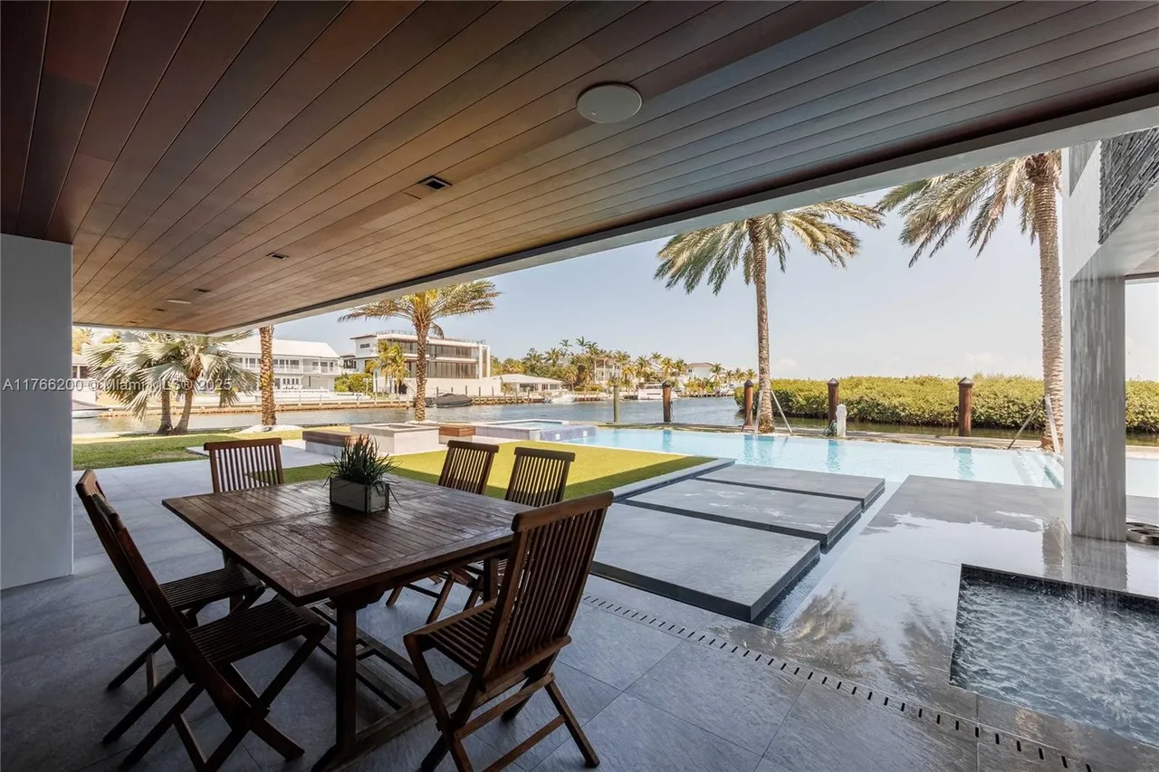 This image showcases a luxurious outdoor patio area with a wooden dining set under a covered ceiling, overlooking a stunning pool and waterfront view. The patio features sleek gray tiling, while the pool area includes modern concrete stepping stones and lush landscaping. Palm trees and waterfront homes are visible in the background, creating a serene and upscale ambiance.