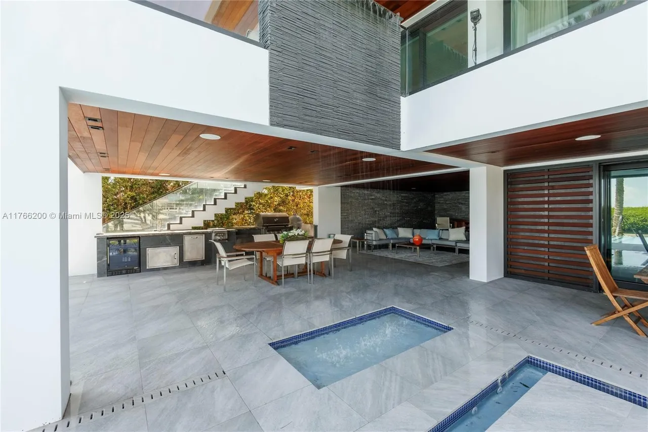 This is a rear exterior view of a modern patio/deck area. The space features a built-in outdoor kitchen, a dining area with a wooden table and chairs, and a lounge area with comfortable seating. There are also unique water features integrated into the patio floor, adding a touch of luxury and tranquility.