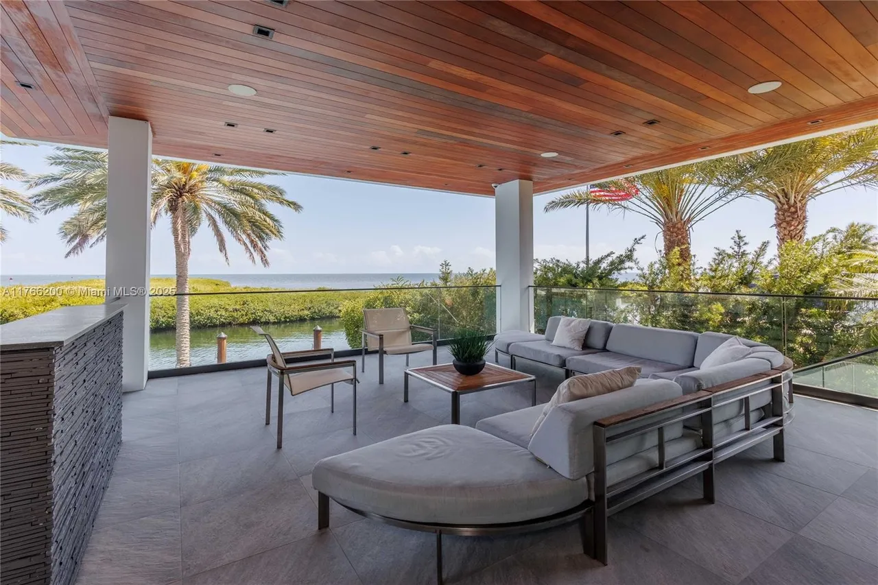 This image showcases a luxurious outdoor patio or balcony area with a stunning waterfront view. The space is furnished with modern outdoor furniture, including a sectional sofa and chairs, arranged around a coffee table. The patio features a sleek design with a wooden ceiling, tiled flooring, glass railings, and white support columns, creating an elegant and inviting atmosphere for relaxation and entertainment.