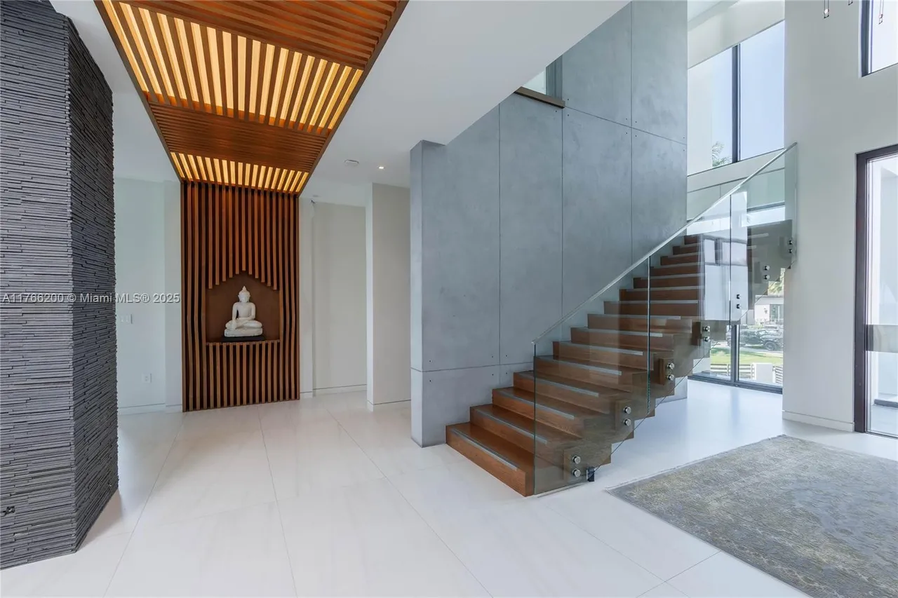 This interior shot showcases a modern hallway with a striking architectural design. A wooden staircase with glass railings ascends alongside a textured gray wall, complemented by a unique wooden ceiling feature with integrated lighting. The space is bright and open, emphasizing clean lines and contemporary style.