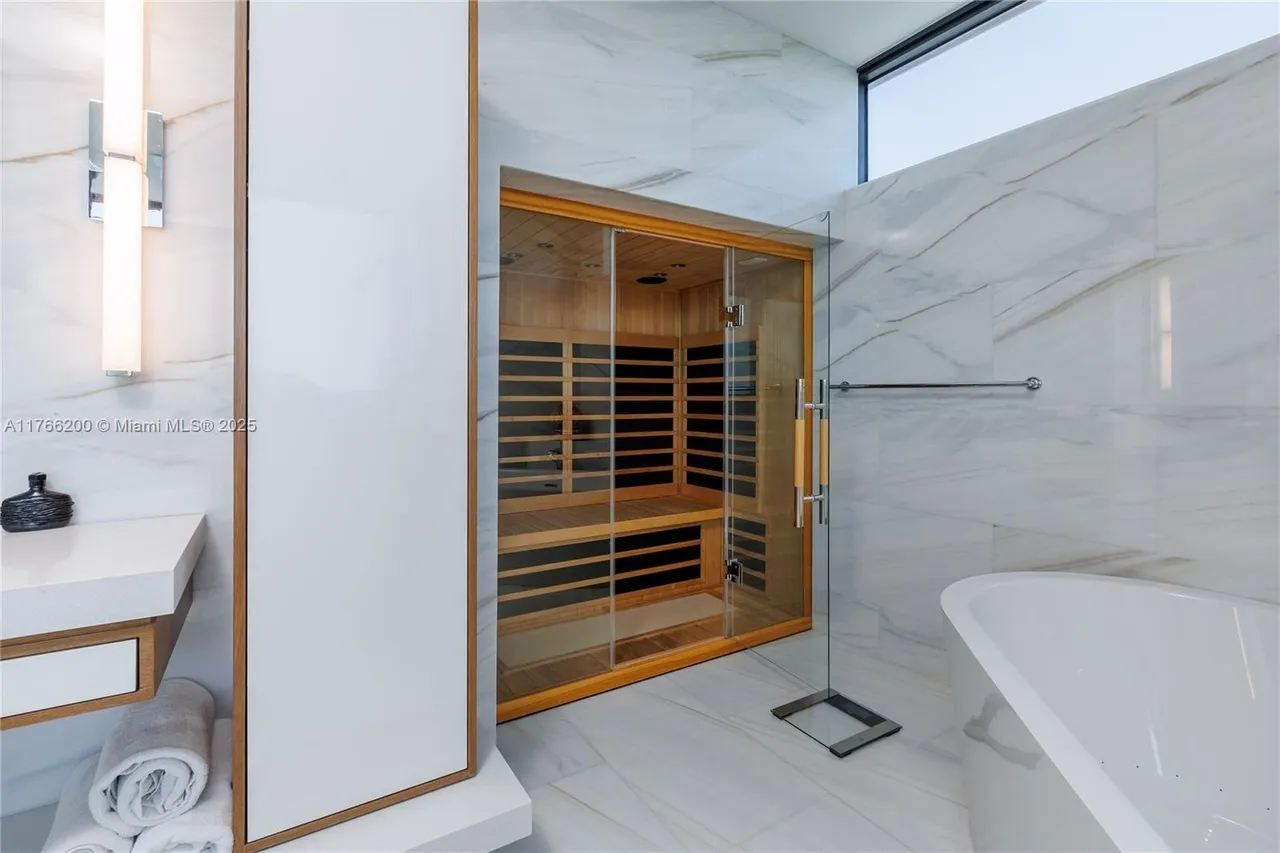 This is a luxurious primary bathroom featuring a modern sauna with a glass door and wooden interior. The bathroom is adorned with marble walls and flooring, complemented by a sleek white bathtub and vanity. The design emphasizes clean lines and a spa-like atmosphere, creating a serene and upscale environment.