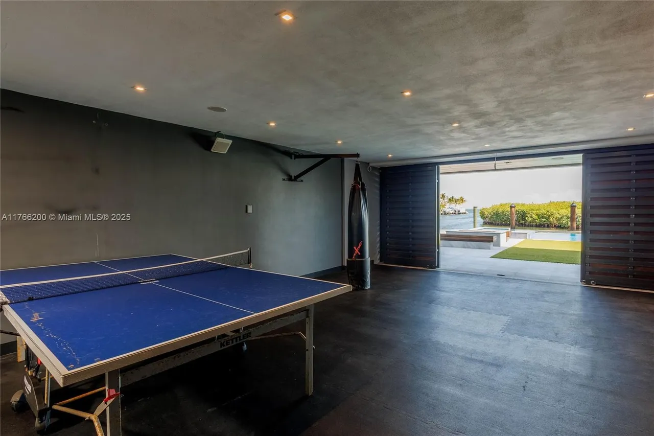 This is an interior shot of a home gym featuring a ping pong table and a punching bag. The room has dark walls and flooring, with recessed lighting in the ceiling. Large sliding doors open to an outdoor area with a pool and waterfront view, creating a seamless indoor-outdoor transition.