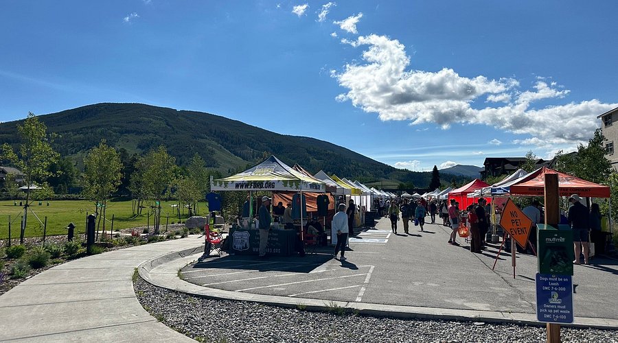This image displays a vibrant outdoor community event or farmers market hosted in a paved area adjacent to a park and mountainous landscape. The scene features several vendor tents arranged in a row, with attendees walking through the space on a bright, sunny day. The location offers a picturesque backdrop with proximity to natural green space, highlighting the active, communal lifestyle of the neighborhood.