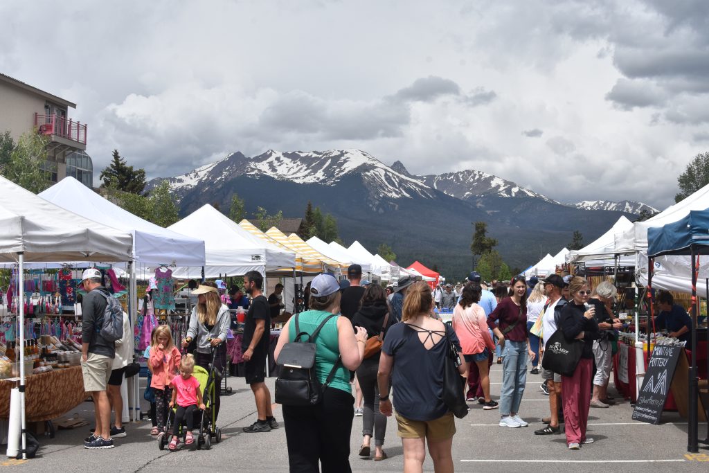 This image captures a vibrant community outdoor market set against the scenic backdrop of snow-capped mountains. The bustling scene features numerous white vendor tents lining an open-air pedestrian area, showcasing a lively atmosphere perfect for local residents and tourists. It highlights the walkability and active lifestyle opportunities available within this neighborhood.