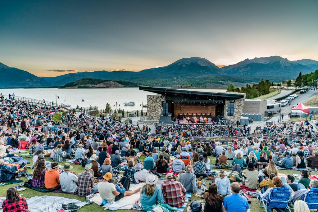 This image showcases a vibrant outdoor community amphitheater situated along a scenic lakefront, offering a premier venue for live performances. The venue features a well-designed stage building with natural stone accents and tiered grassy lawn seating that provides excellent views of the mountains in the background. It represents an exceptional recreational amenity, highlighting a lifestyle centered around culture and natural beauty.